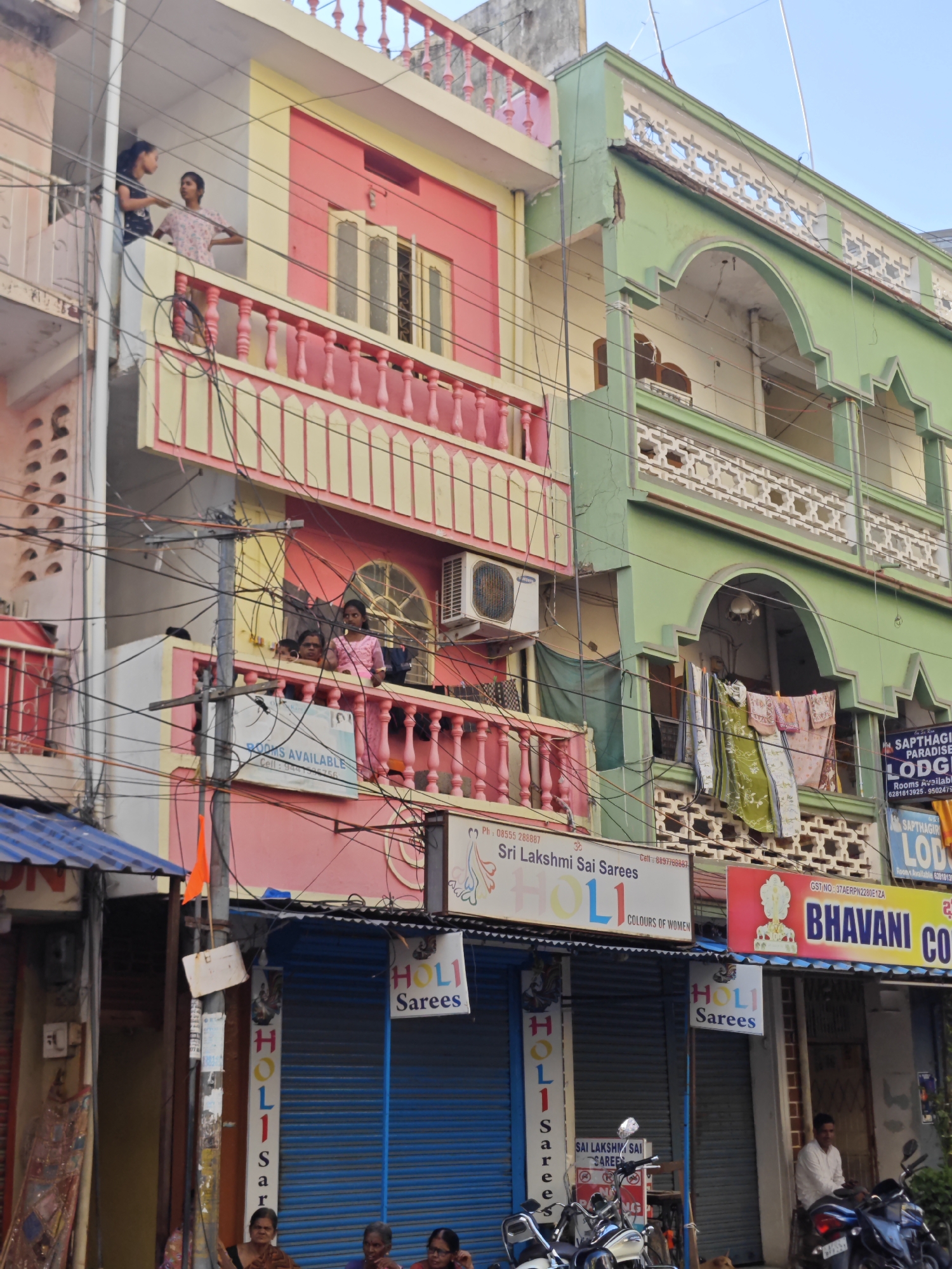 This image captures a street scene in Puttaparthi, India, focusing on the colorful architecture of multi-story buildings. The foreground features the facades of shops with blue roller shutters. One shop displays a sign for "HOLI Sarees" with a colorful logo. To the right, another shop has a prominent sign for "BHAVANI CONFECTIONERY." A man in a white shirt is seated on a motorcycle in front of this shop, while another motorcycle is parked in front of the "HOLI Sarees" shop.
Above the shops, balconies are visible. On the pink and yellow building, two young women are standing on a balcony, with another person visible behind them. A sign on this balcony reads "ROOMS AVAILABLE." The green building to the right has clothes hanging from a balcony. Laundry lines are also visible on another balcony. The overall environment appears to be a commercial street with a mix of residential and business establishments. Power lines crisscross the scene, indicating a busy urban setting. The sky is visible in the upper portion of the image.