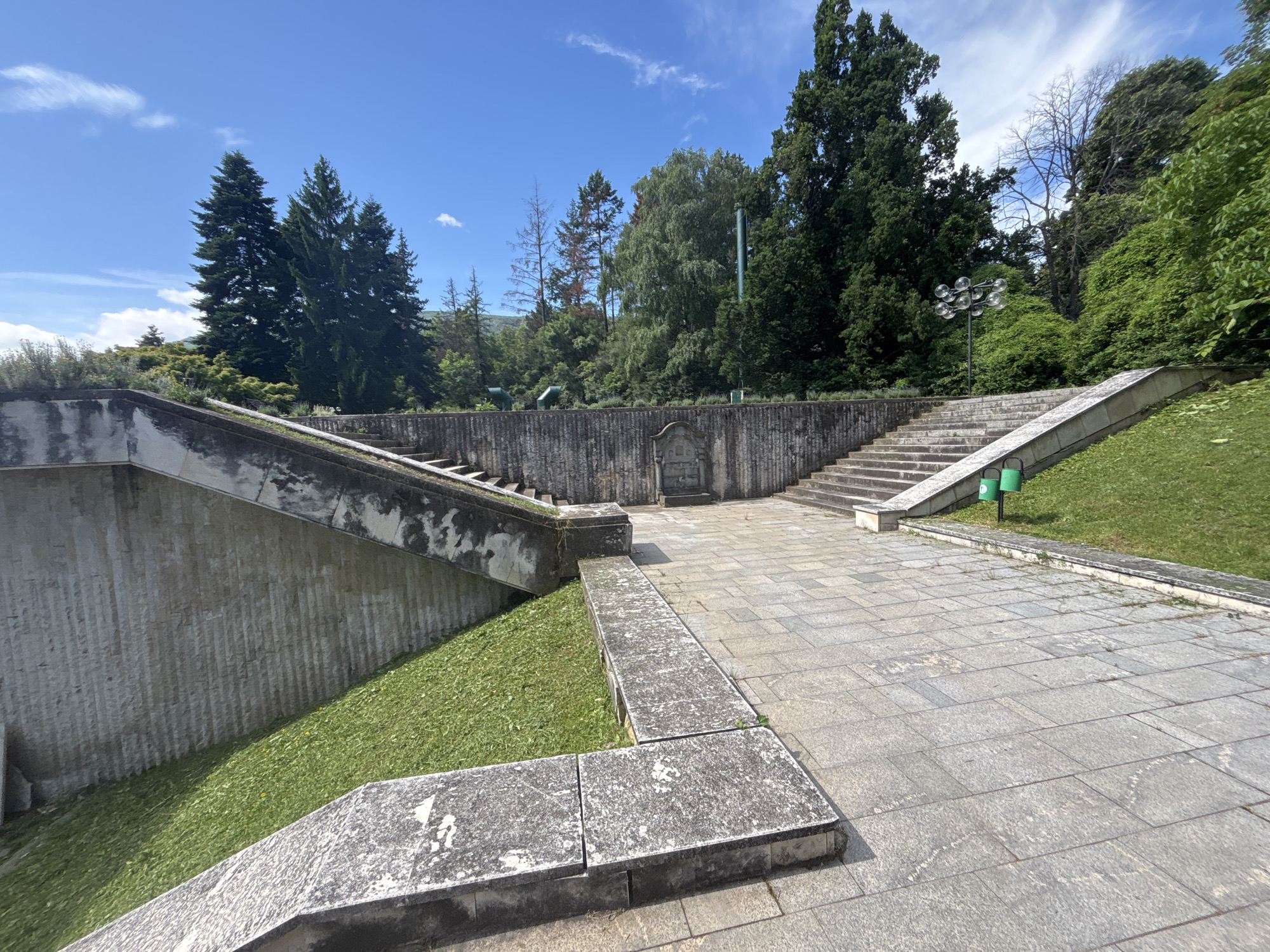 The image depicts an outdoor stone staircase and paved area with lush green grass and trees under a bright blue sky with scattered clouds. The architecture appears to be a public space, possibly a park or garden, with concrete and stone elements forming the steps and retaining walls. There are two green trash receptacles visible near the base of the stairs. The scene is devoid of people. Based on the visual cues, this location is likely the Borisova Gradina park in Sofia, Bulgaria, specifically the area around the National Palace of Culture (NDK) gardens, known for its terraced design and modern public art installations.