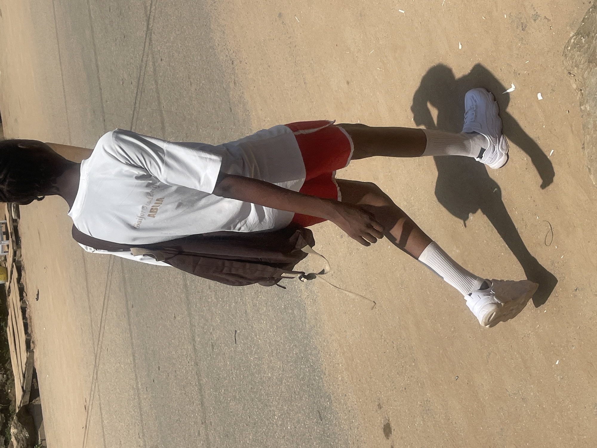 A person wearing a white t-shirt with "Baifor Academy ABUJA" printed on it, red shorts with white trim, white socks, and white sneakers walks on a dusty road. They are carrying a dark brown backpack over their right shoulder. The sun casts a prominent shadow of the person on the ground. In the background, a wooden structure with planks and some yellow objects are visible, suggesting an urban or semi-urban environment within Abuja, Nigeria. The road appears to be made of asphalt and dirt.