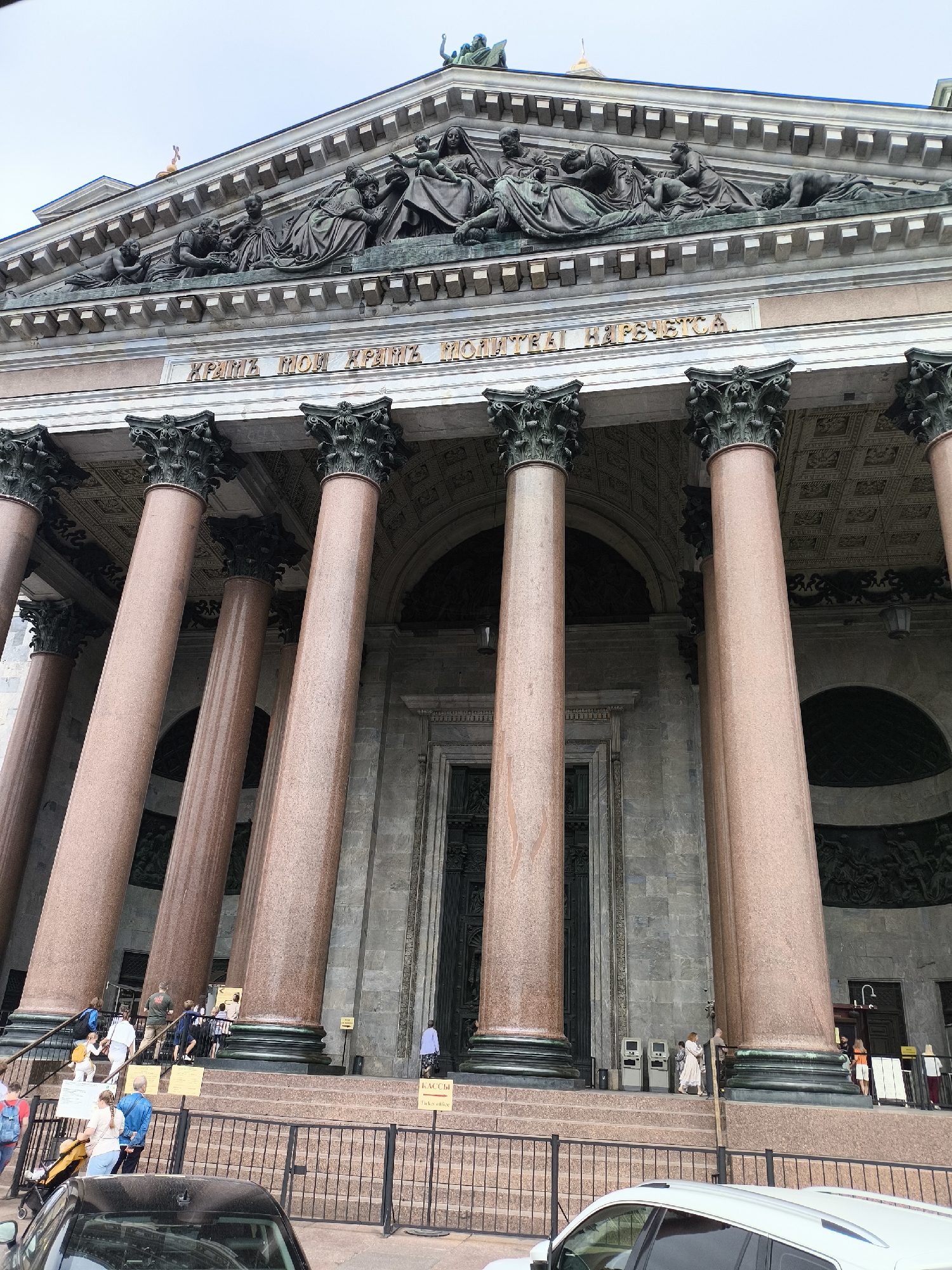 The image captures a low-angle, full-frame shot of the imposing facade of St. Isaac's Cathedral in Saint Petersburg, Russia. The foreground is dominated by several massive pink granite columns with ornate, dark green capitals. Above the columns, a frieze with sculpted figures is visible, and further up, the pediment features more elaborate sculptural reliefs. A golden inscription, likely in Russian Cyrillic, is set into the entablature between the frieze and the capitals.
At the base of the cathedral, a wide set of stairs leads up to a grand entrance. Several people are on the stairs and in front of the entrance, some ascending, some descending, and others standing. A black railing separates the stairs from the sidewalk area in front of the building. A black car is parked in the lower left corner of the frame, and a white SUV is partially visible in the lower right. The overall atmosphere is one of grandeur and historical significance, with a steady flow of visitors interacting with the iconic landmark. The sky above is overcast, suggesting a temperate climate.