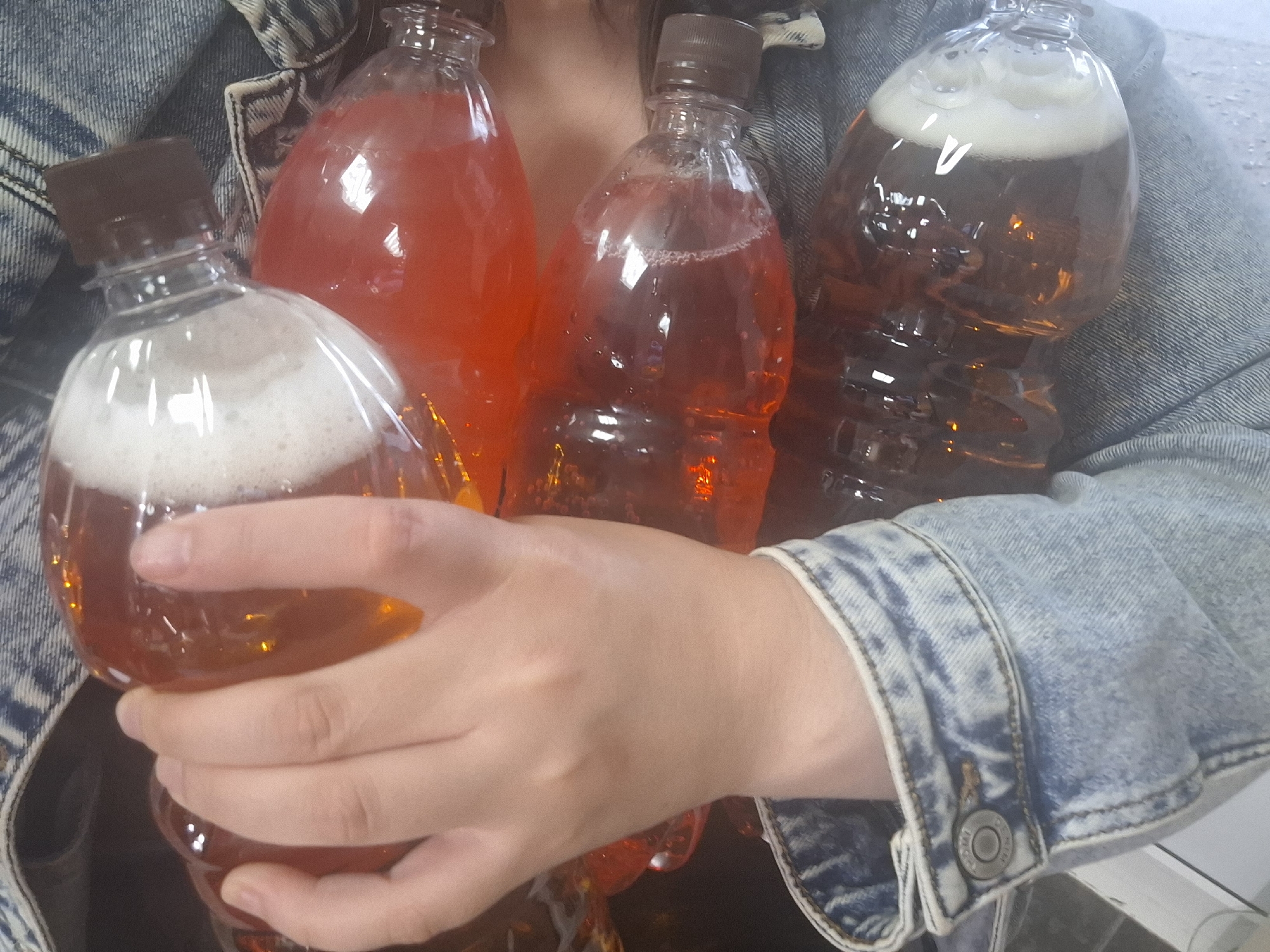 A person wearing a denim jacket is holding four plastic bottles filled with liquids. Two bottles contain a red or orange liquid, while the other two hold a brown liquid with a foamy white head. The scene provides no evidence regarding the city of Chaikovsky, Russia.