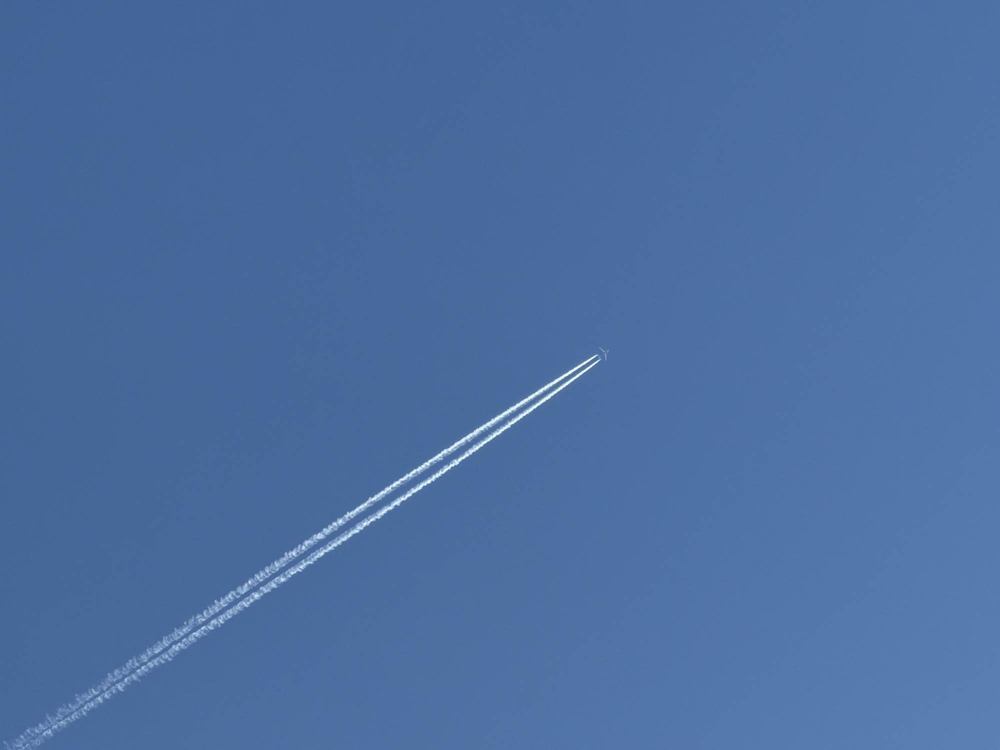 The image displays a clear, cloudless blue sky, observed from Izhevsk, Russia. A passenger aircraft is visible in the lower right quadrant, silhouetted against the bright sky, generating a prominent white contrail. This contrail, formed of two distinct, parallel vapor trails, extends diagonally across the frame from the upper left corner downwards towards the aircraft. The aircraft appears to be traveling from the upper left to the lower right of the frame, leaving the dual contrail in its wake. No other objects, landscapes, or individuals are present in the scene.