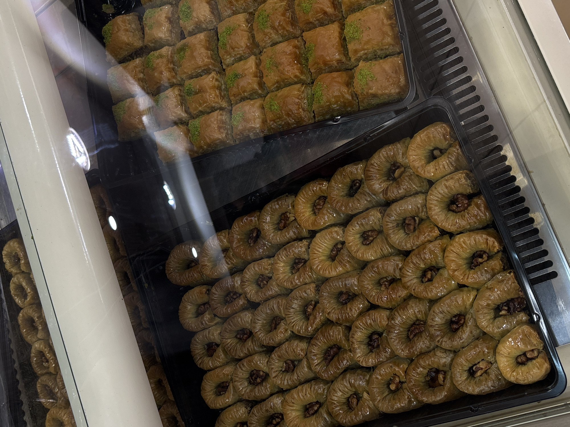 The image shows two trays of baklava pastries in a display case. The top tray contains square pieces of baklava, each topped with green pistachio. The bottom tray features round, spiral-shaped baklava with a walnut in the center of each piece. The display case is likely located in a dessert shop or bakery in Perm, Russia.