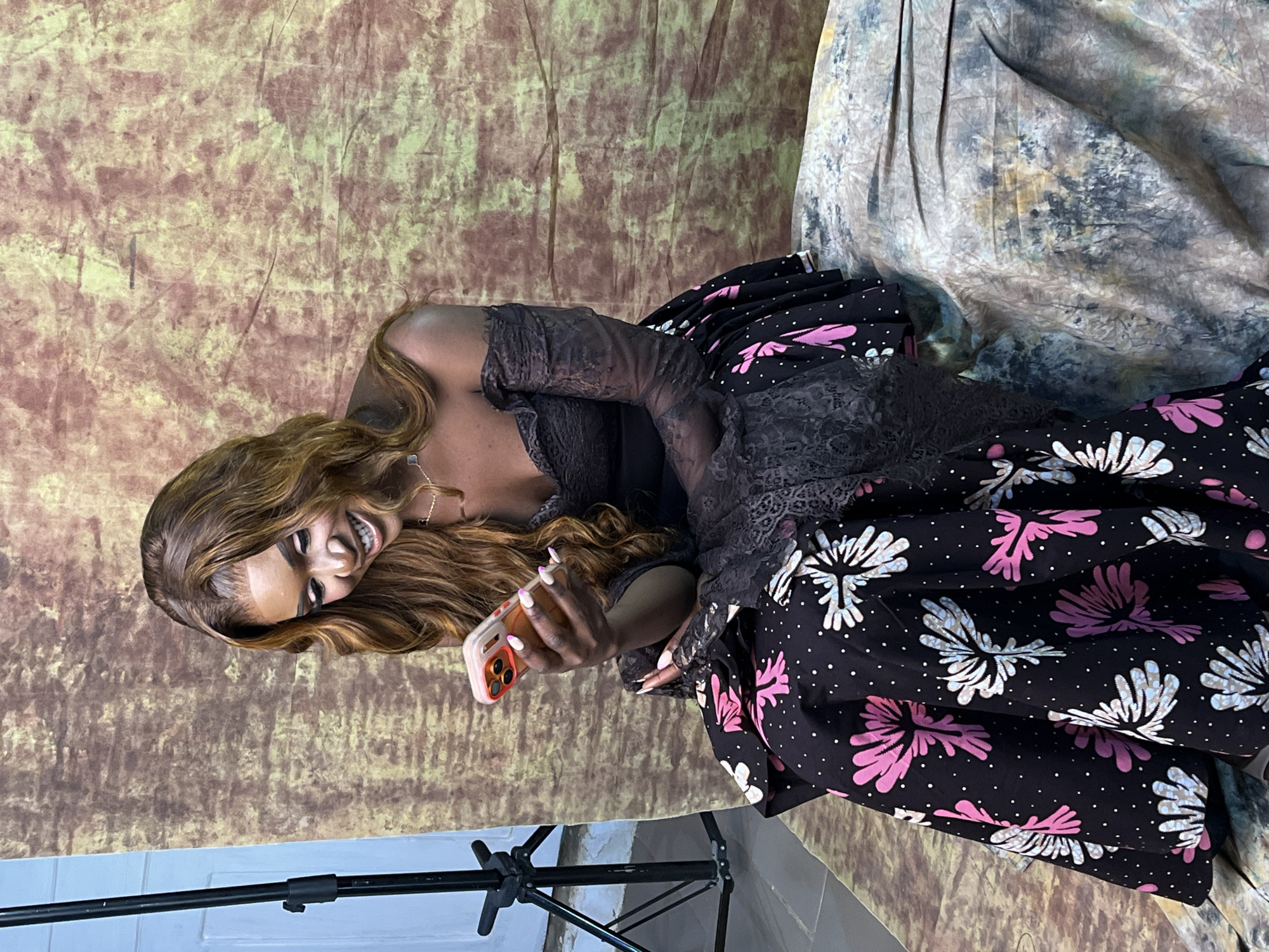 A woman is holding a phone, smiling and looking at the phone. She is wearing a black lacy dress with a black skirt with white and pink floral designs. She has brown hair and is wearing a gold necklace. There is a backdrop with a mottled, earthy tone and another backdrop with a marbled effect. A photography light stand is visible in the bottom left corner. The location is implied to be Barakin Akawo, Nigeria.