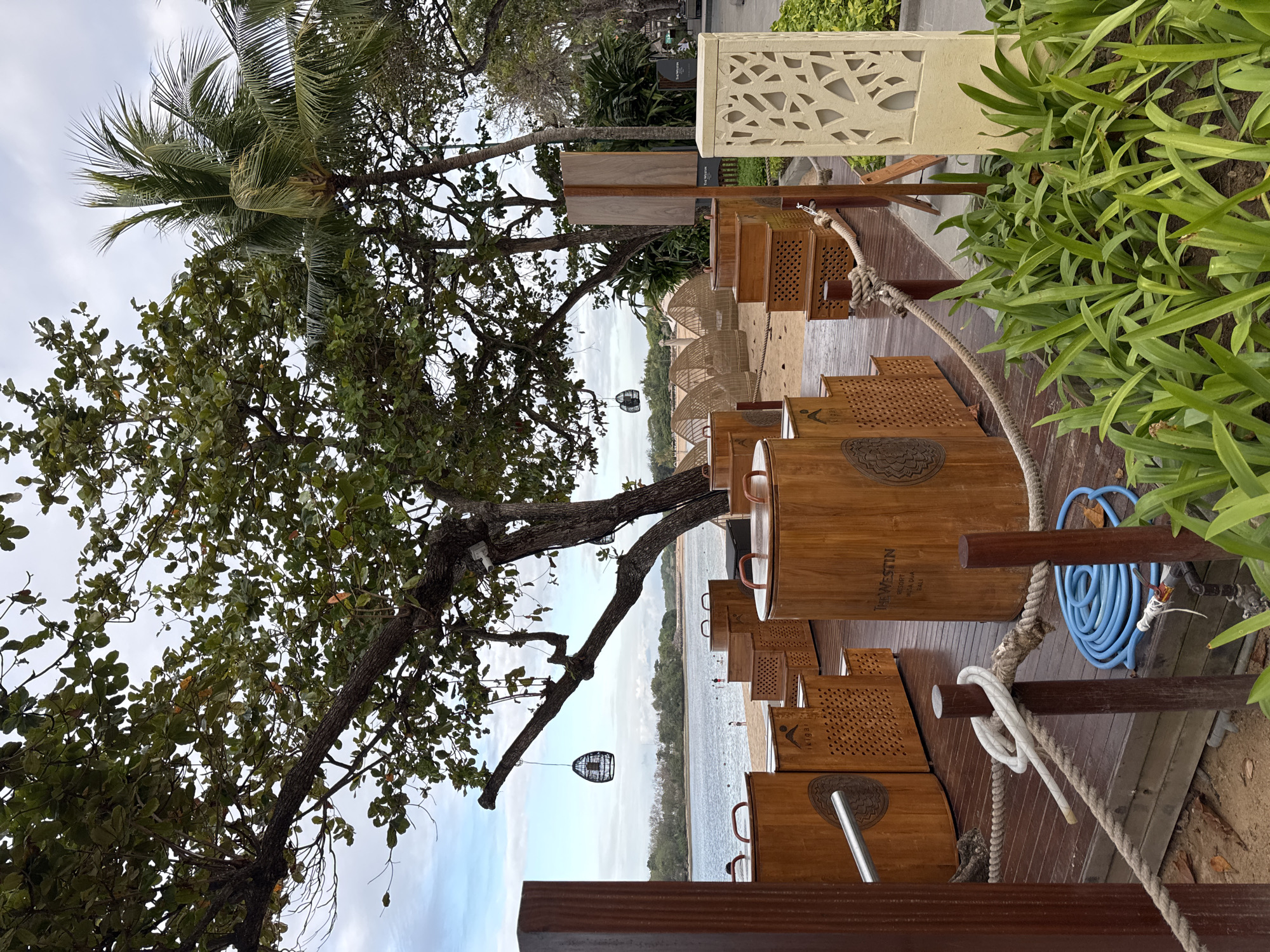 An outdoor seating area at a resort in Kuta Selatan, Indonesia, is depicted in this medium-angle, eye-level shot. The foreground features a wooden deck with a rope barrier, a coiled blue hose, and several large, cylindrical wooden containers. The containers have handles and some have wooden lids. The text "The Westin RESORT NUSA DUA BALI" is visible on one of the containers. Behind the deck, a sandy beach leads to calm water with a few people visible in the distance. Lush green trees, including a large tree with intricate branches, dominate the upper portion of the frame, with palm trees to the right. The sky is overcast with a few patches of blue. In the background, thatched-roof cabanas or seating structures are arranged on the sand. A light-colored, decorative pillar with a cut-out pattern stands on the right side of the deck. The overall scene suggests a resort setting by the sea.