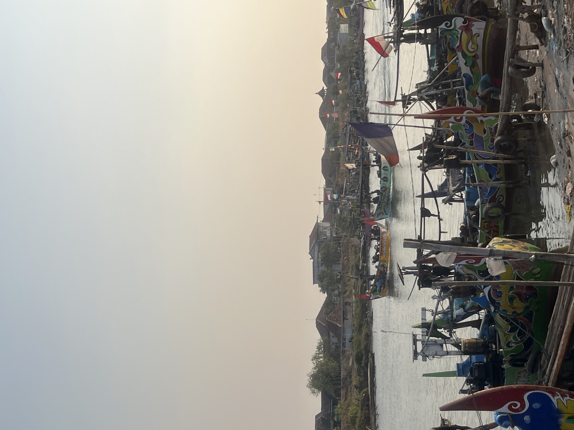 This is a medium shot, taken outdoors, of a fleet of brightly colored fishing boats moored on a riverbank in Kedungmalang, Indonesia. The sky is a pale, hazy blue, transitioning to a warm yellow at the horizon, indicating either dawn or dusk. In the background, a line of buildings with terracotta roofs and some trees form a backdrop along the river's edge.
The foreground and midground are dominated by the fishing boats. These boats are of varying sizes and appear to be made of wood. Many are ornately decorated with vibrant patterns, featuring swirling lines, floral motifs, and what appear to be stylized animal figures in yellow, green, blue, red, and white. Some boats have multiple people visible, either standing or sitting. Small flags, including what appears to be the French flag (tricolor of blue, white, and red) and Indonesian flags (red and white), are flown from some of the boats and poles.
The river itself is calm, with ripples reflecting the light. The riverbank in the immediate foreground is a mix of muddy earth and debris, including discarded tires and plastic waste. Several wooden posts and structures are visible along the bank, suggesting a functional docking area. The lighting is soft and diffused, casting gentle shadows. The overall impression is a bustling, colorful scene of a traditional fishing community.