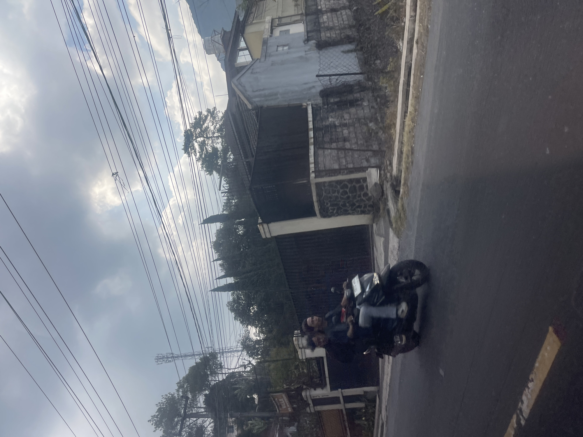 In Ciloto, Indonesia, on a paved road, two individuals are riding a black motorcycle. The person in front is operating the vehicle, while the person behind is seated. They are traveling from left to right in the frame. On the left side of the road, a gate with a sign that reads "VILLA" is visible, along with trees and a house. On the right side of the road, a fence and a multi-story building are present. The sky above is cloudy.