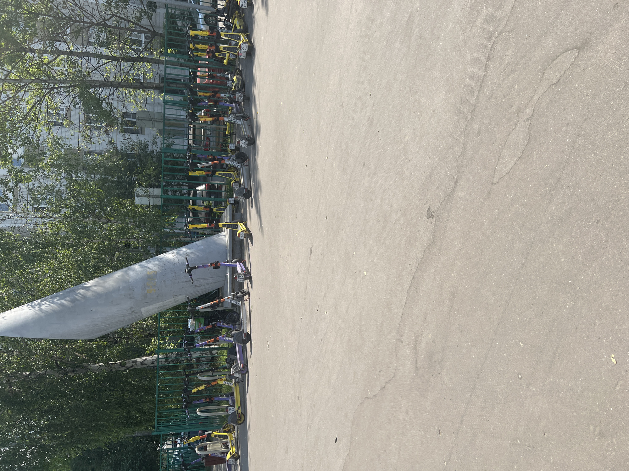 The image depicts a row of parked electric scooters along a green metal fence. The scooters are of various colors, including yellow, purple, and gray, and many have identification numbers visible on them. Behind the fence, a tall, grey, conical monument is visible, tilted diagonally. Trees with green foliage are positioned around the monument and the fence. In the background, a multi-story building with numerous windows can be seen. The scooters are parked on a paved surface. The scene appears to be a public area in Moscow, Russia, likely a designated parking zone for shared electric scooters.