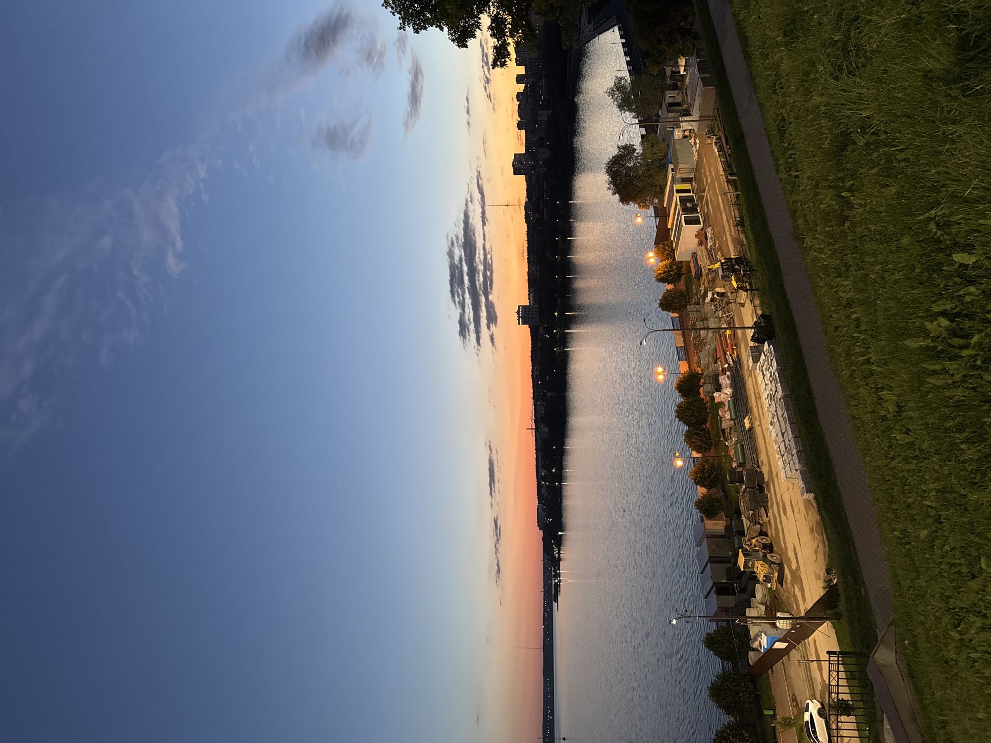 The image depicts a sunset over the Izh River in Izhevsk, Russia. The sky displays a gradient of blue and orange hues. A cityscape lines the opposite bank of the river, with silhouetted buildings and a prominent radio tower. The river itself is calm and reflects the colors of the sky. In the foreground, a grassy embankment slopes down towards a waterfront area. This area appears to be under construction, with piles of materials, construction equipment including a yellow bulldozer, and modular buildings visible. A paved pathway runs along the construction site, and streetlights illuminate the scene. A white car is parked on the left side of the pathway.