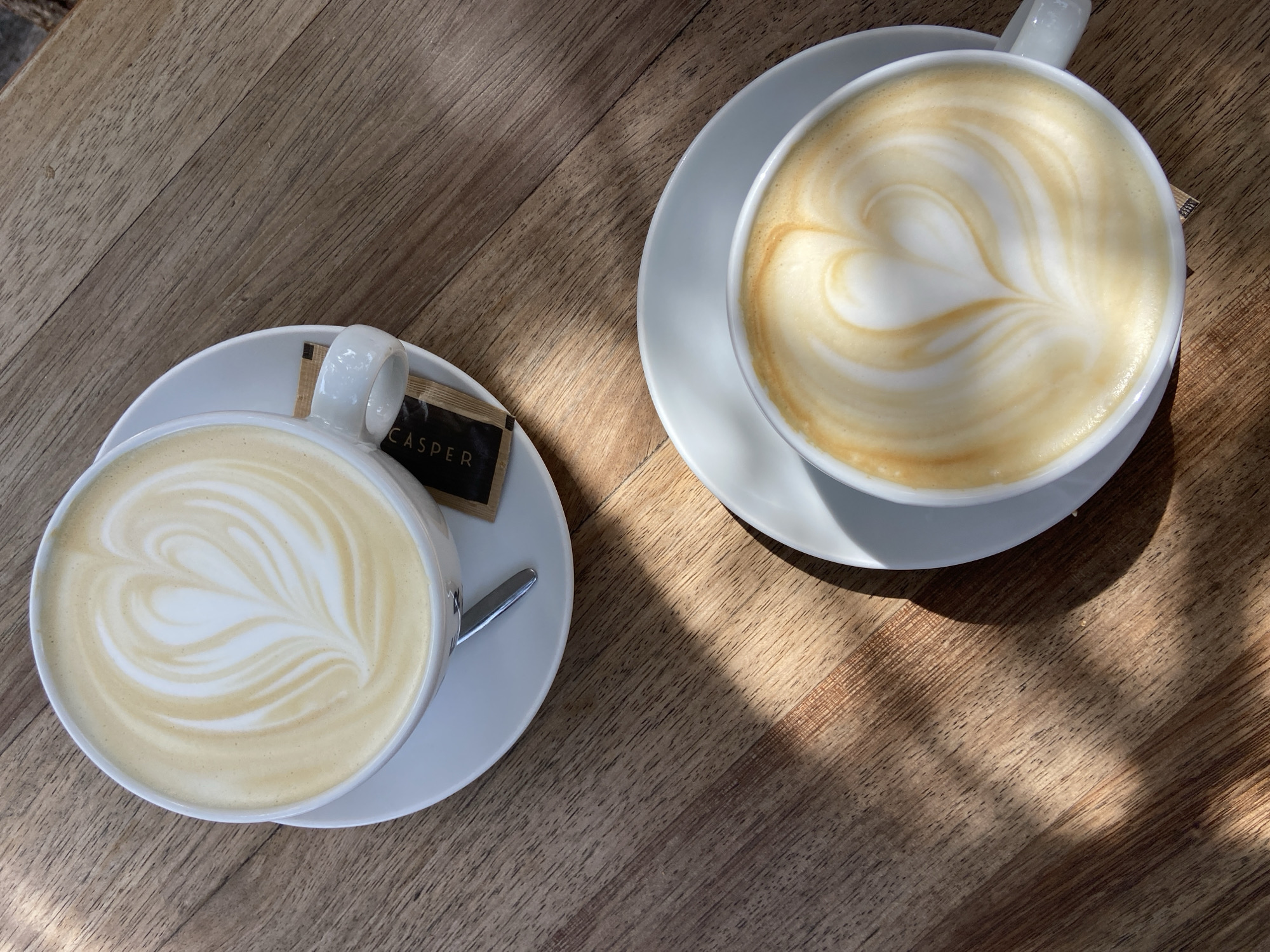 This overhead shot captures two artfully prepared lattes, each featuring intricate heart-shaped latte art, served in white cups on matching saucers. The beverages are placed on a warm, light brown wooden table, suggesting a comfortable cafe setting in Budva, Montenegro.
One cup is positioned in the upper right, its latte art forming a soft, well-defined heart. The other cup, in the lower left, also displays a heart design in its foam, along with additional swirled patterns. A silver spoon rests on the saucer of the lower-left cup. Beside this cup, a small, dark brown rectangular packet, likely containing sugar or sweetener, is visible, with the word "CASPER" printed prominently in white capital letters.
The wooden table surface exhibits a distinct grain, adding to the natural aesthetic. Strong, elongated shadows stretch across the table from the top right towards the bottom left, indicating bright sunlight and suggesting either an early morning or late afternoon. The clear contrast between the sunlit and shadowed areas creates a dynamic visual effect, enhancing the serene and inviting atmosphere of a relaxing coffee break.