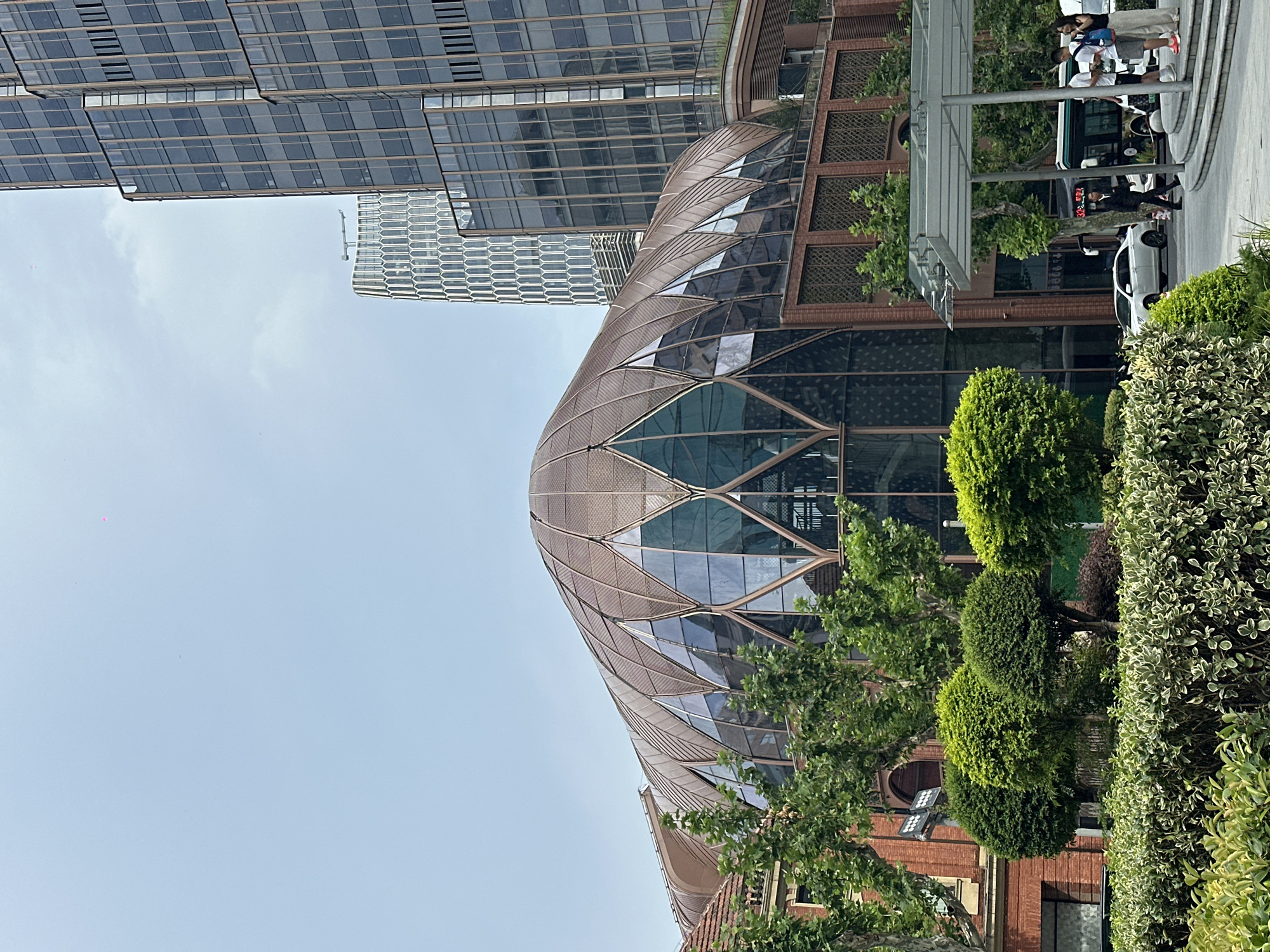 The image depicts the exterior of a unique modern building with a curvilinear, petal-like glass and metal roof, likely a commercial or cultural space, situated in Shanghai, China. The building is surrounded by lush greenery and is adjacent to taller, contemporary skyscrapers with glass facades. In the foreground, a well-maintained hedge and neatly trimmed bushes are visible. A street scene unfolds to the right, featuring a white sedan, a bus, and several individuals walking. One man in a suit is walking towards the left, while a group of three people, two men and a woman, are standing on a set of steps, with one woman appearing to be holding a phone. The sky is overcast.