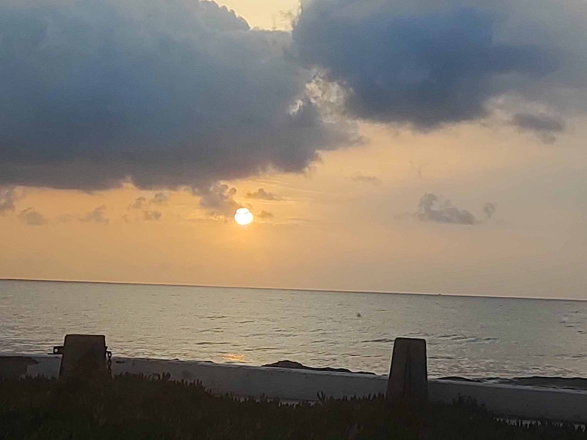 The image depicts a serene sunset over the ocean, presumably at Playa, Spain. The sun, a bright orb, is partially obscured by dark, dramatic clouds, casting a warm, golden glow across the sky and water. The ocean is calm, with gentle ripples reflecting the soft light. In the foreground, the dark silhouettes of dune vegetation and two concrete bollards suggest a beach setting. The overall atmosphere is one of tranquility and natural beauty.
