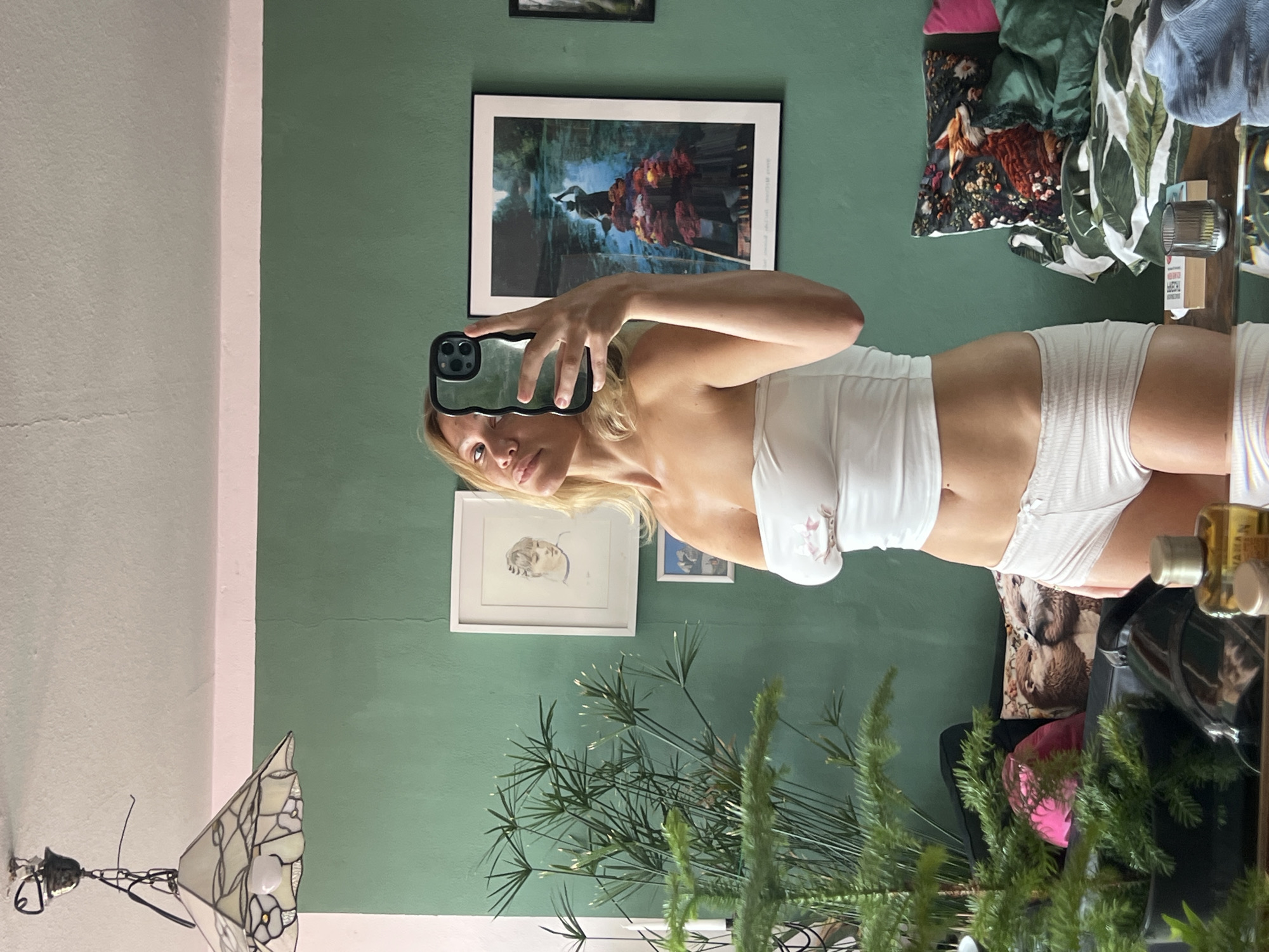 The image shows a woman taking a mirror selfie in a room. She is wearing a white strapless top and white shorts. She is holding a black phone in her right hand. The background shows a green wall with several framed pictures, a plant, and a bed with pillows and bedding. A lamp hangs from the ceiling. The room appears to be a bedroom or a living room. The location of Mühldorf a. Inn, Germany is not indicated in the image.