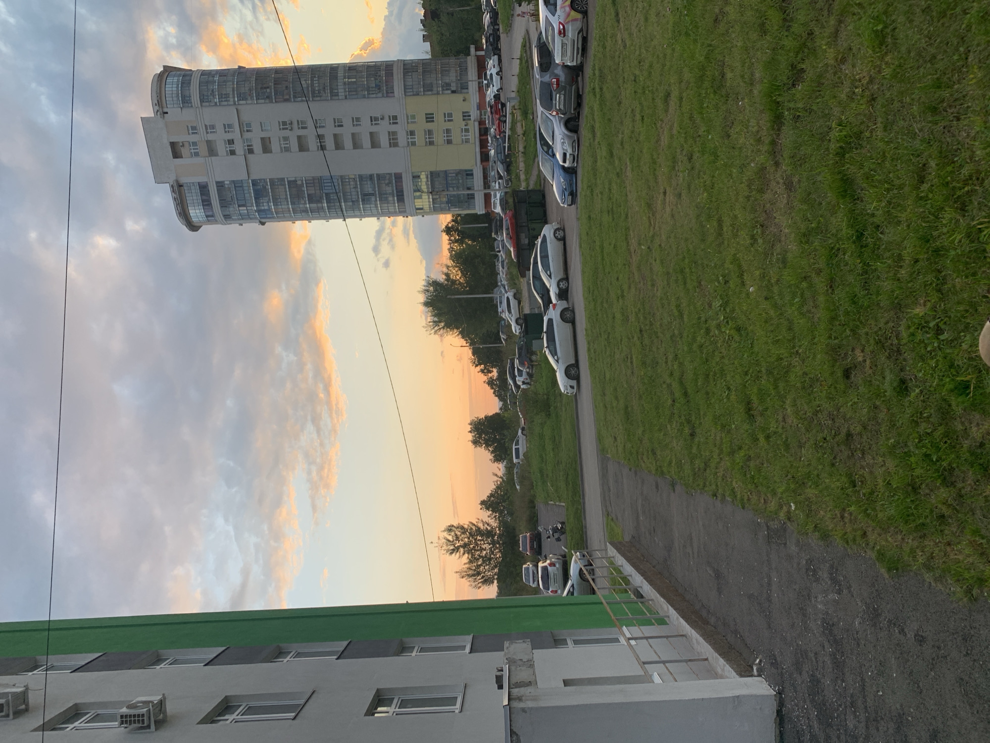 The image depicts a scene in a residential area in Ufa, Russia, during sunset. A modern, multi-story apartment building with a curved glass facade dominates the background. In the foreground, a smaller, light-colored apartment building with a green vertical accent is visible on the left. A parking lot filled with numerous cars is situated between the two buildings, extending towards the horizon. The sky is filled with dramatic clouds illuminated by the setting sun, casting warm hues of orange and pink. A patch of green grass is in the middle ground, separating the paved walkway from the parking lot. A few trees are scattered in the distance, adding to the urban landscape. The scene appears peaceful and mundane, characteristic of a residential neighborhood.