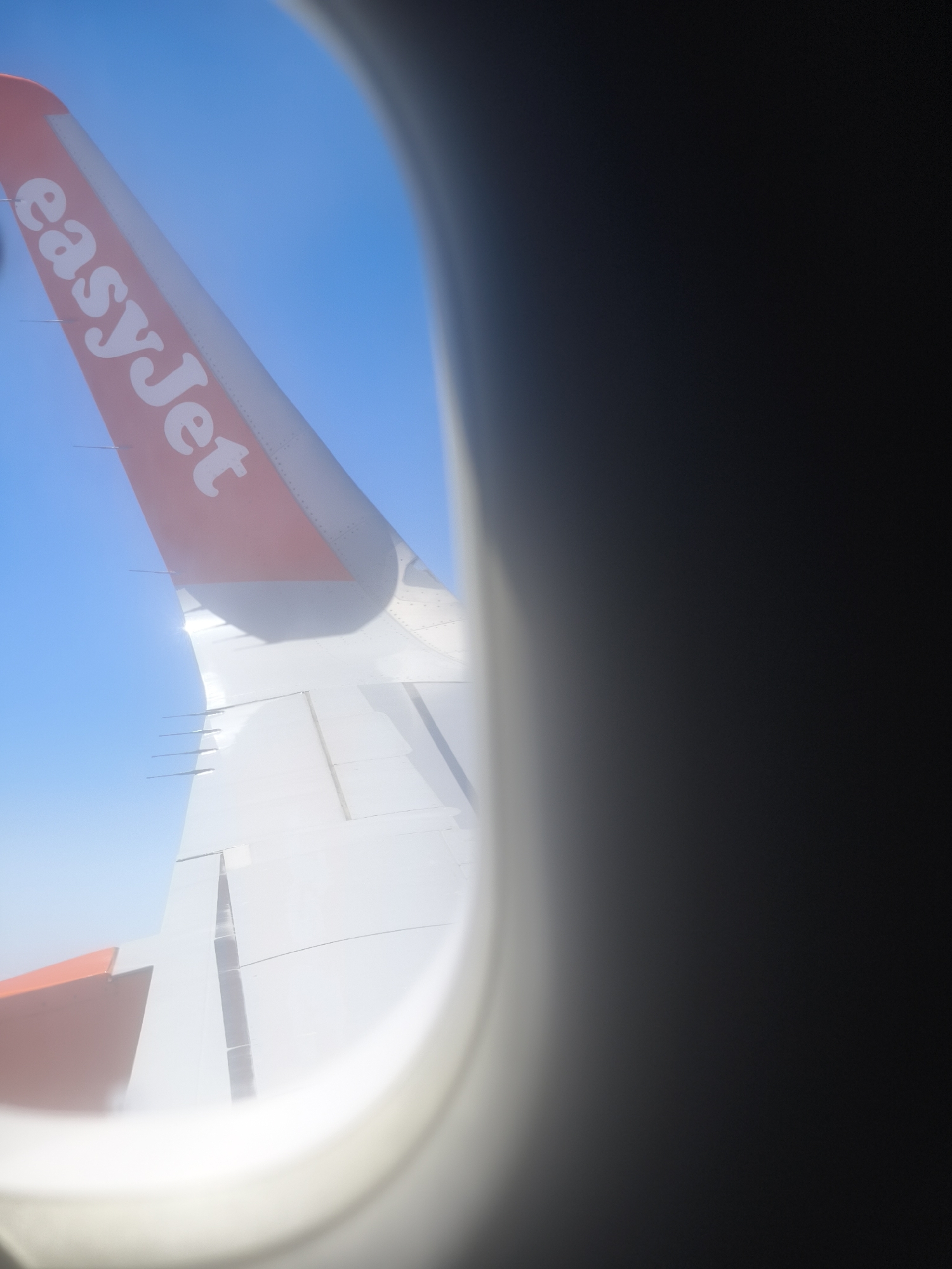 The image displays an aerial view of an EasyJet airplane wing seen through an aircraft window. The wing is predominantly white with a bright orange tip featuring the "easyJet" logo in white lettering. The wing's surface shows panel lines and several small vertical fins, indicative of its aerodynamic structure. The background is a clear blue sky, suggesting fair weather. The right side of the image is dominated by the dark, blurred interior of the aircraft cabin, likely the window frame and surrounding structure, with a slight reflection or fogging on the window. The perspective is from inside the plane, looking out towards the wing and sky. There is no discernible location in the city of Trebbin, Germany.