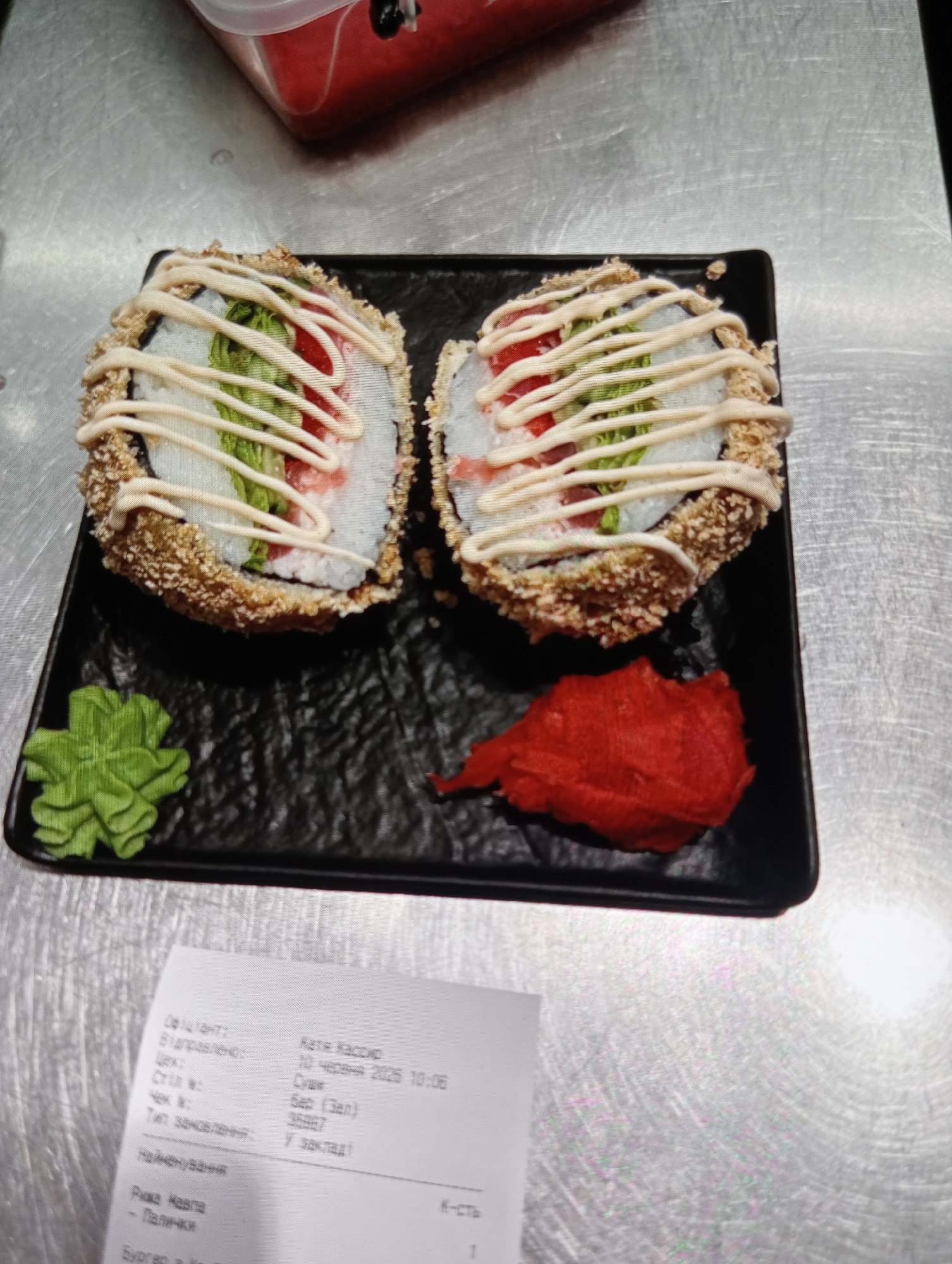 Two pieces of sushi are presented on a black rectangular plate. The sushi pieces are round and have a crispy, fried outer layer. They are topped with a white sauce drizzled in a criss-cross pattern, and a visible green vegetable and red ingredient can be seen inside. To the left of the plate is a dollop of green wasabi, and to the right is a pile of red pickled ginger. In the lower part of the image, a receipt is visible, showing Cyrillic text and indicating a date of "10 червня 2025 10:06." The receipt also lists "Миколаїв" which translates to Mykolaiv, Ukraine. The scene is set on a metallic surface, likely a table.