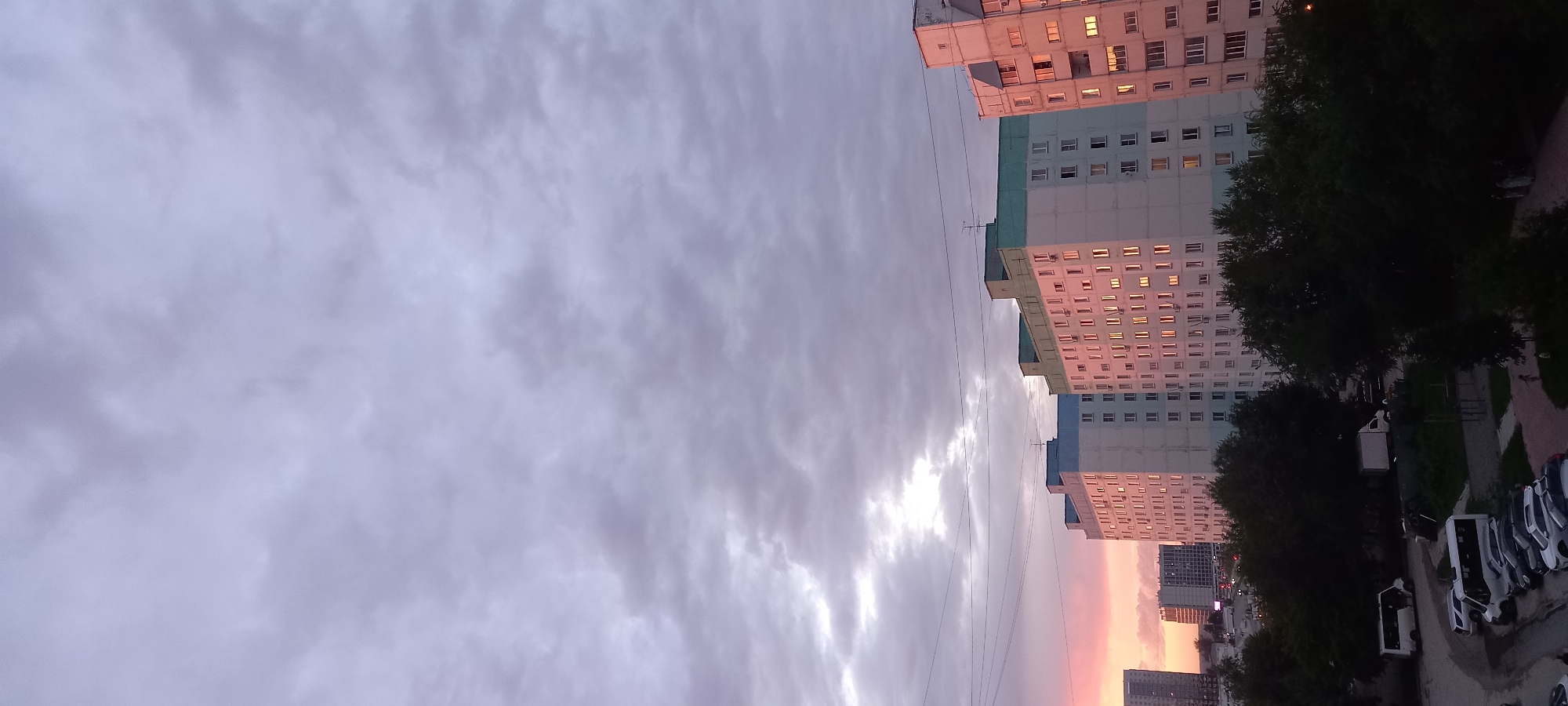 The image shows a residential area in Novosibirsk, Russia, during sunset. The sky is filled with clouds, with a hint of pinkish-orange hues from the setting sun on the left side of the frame. Several apartment buildings are visible, with windows reflecting the warm colors of the sunset. Some of these buildings appear to be older, panel construction, typical of Soviet-era housing, while others might be more modern. Green trees are present in the foreground and middle ground, obscuring parts of the lower floors of the buildings and the street level. Below the buildings, a parking lot is visible with several cars parked, including white vans and passenger vehicles. In the distance, other buildings and possibly streetlights can be seen. The overall scene depicts a typical urban evening in a residential district, characterized by the interplay of man-made structures and natural light. There are no discernible individuals engaged in specific activities, and the focus is on the architecture and the atmospheric conditions of the sunset.