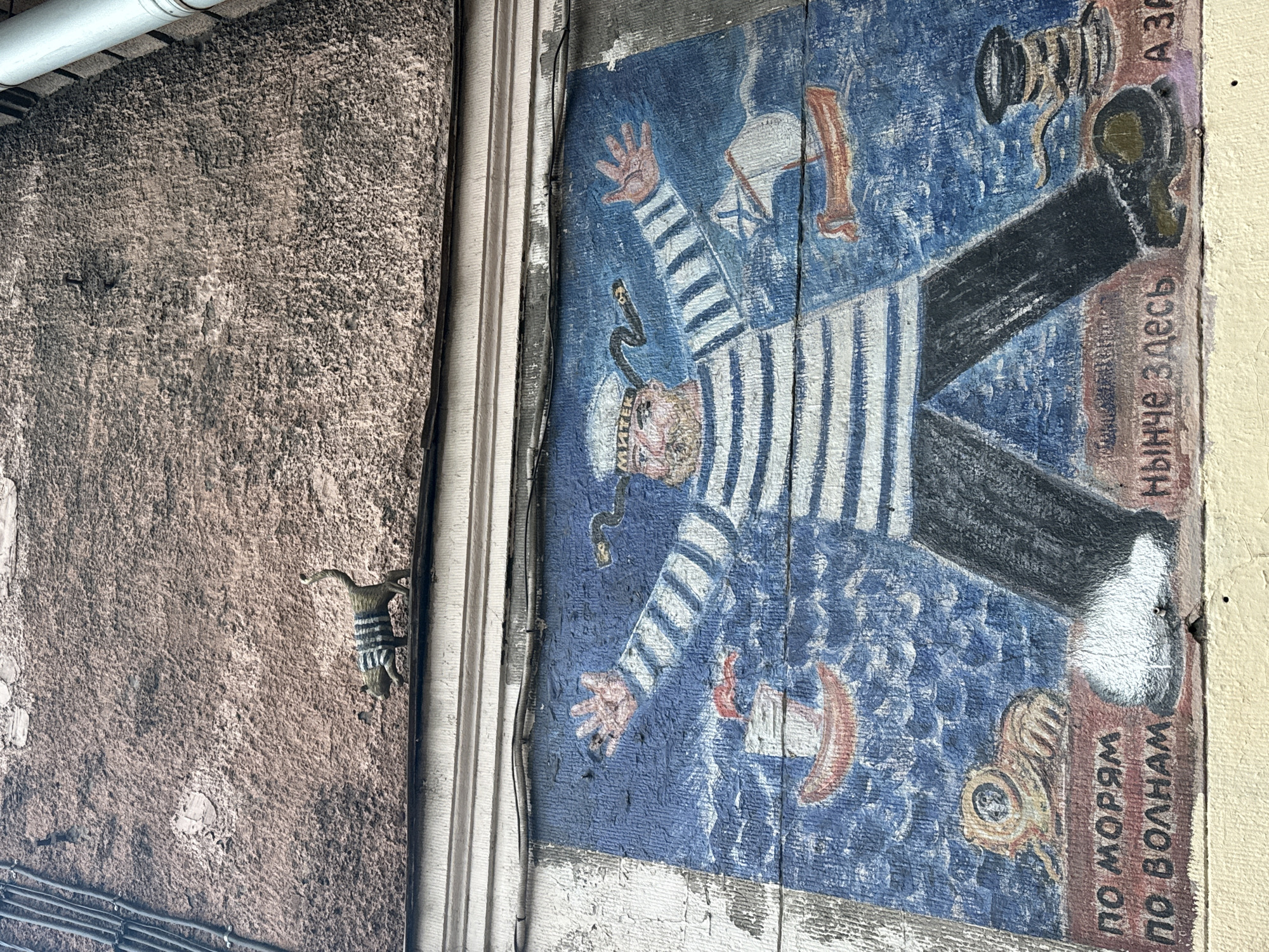 The image shows a mural painted on a wooden wall in Saint Petersburg, Russia. The mural depicts a stylized sailor wearing a striped shirt and hat. A black snake is wrapped around his neck. He is lying on his back with arms and legs outstretched, against a blue background with nautical objects. Russian text is present on the right side. The wall is weathered and peeling. Adjacent to the mural is a rough, brown wall with a small figurine mounted on it.
