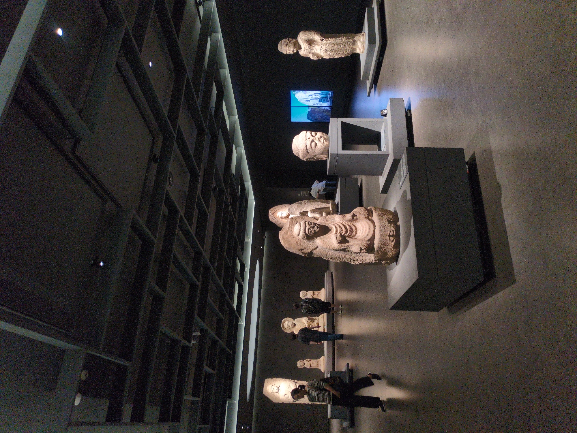 The photograph depicts the interior of the Gyeongju National Museum in Gyeongju-si, South Korea. Displayed are several ancient stone sculptures, possibly sarcophagi or related artifacts, arranged on pedestals and platforms. Some visitors can be seen observing the exhibits. The museum's architecture is visible, including the modern design of the wall with a grid pattern. The lighting is a combination of ambient light and spotlights illuminating the displays.