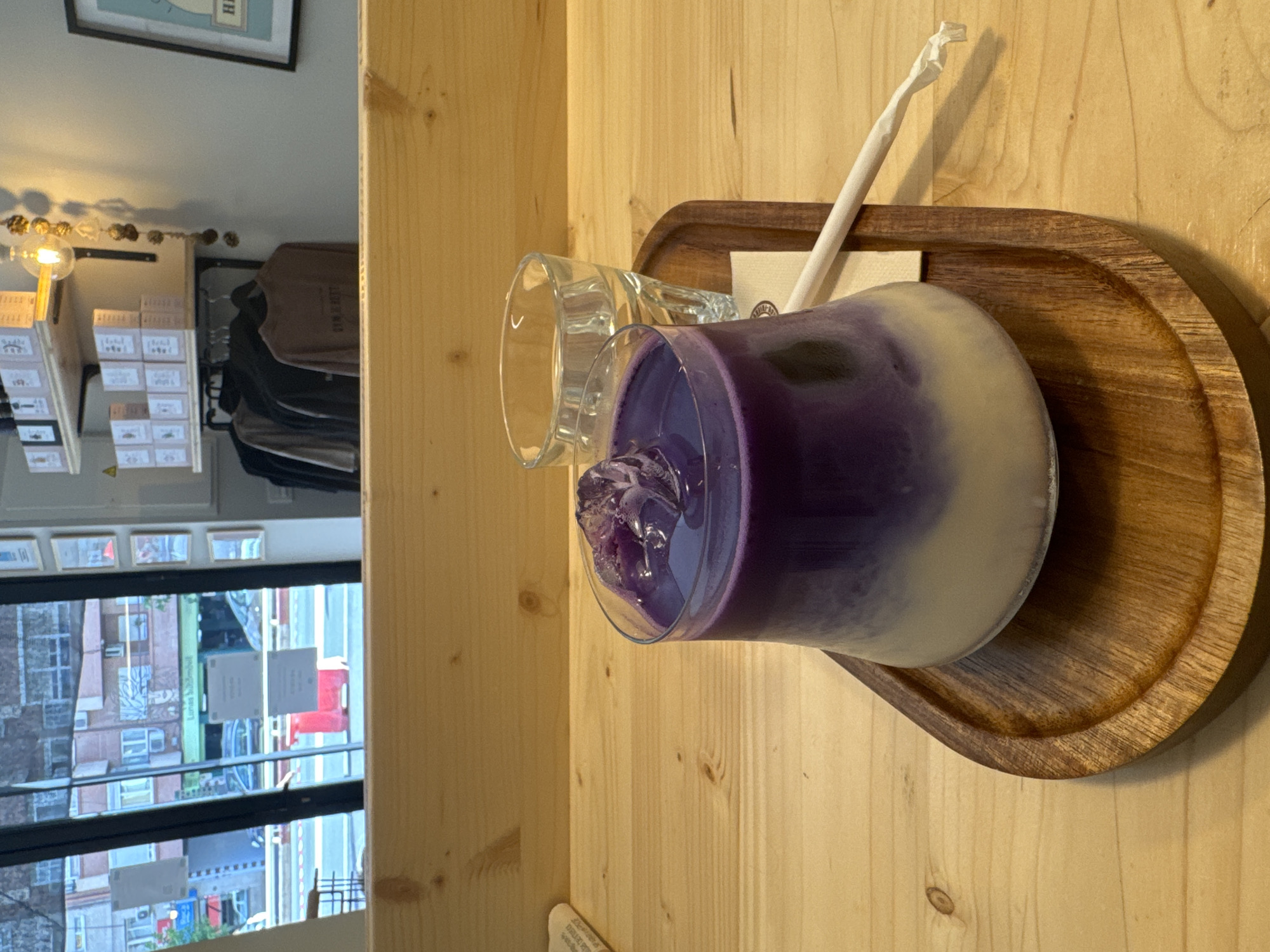 A layered purple and white drink with ice is presented on a wooden tray with a white straw and a glass of water on a wooden counter. Behind the counter, through a large glass window, a street scene in Murcia, Spain, is visible, featuring buildings, cars, and pedestrian traffic. The interior of the establishment shows shelves stocked with boxes and a clothing rack with shirts.