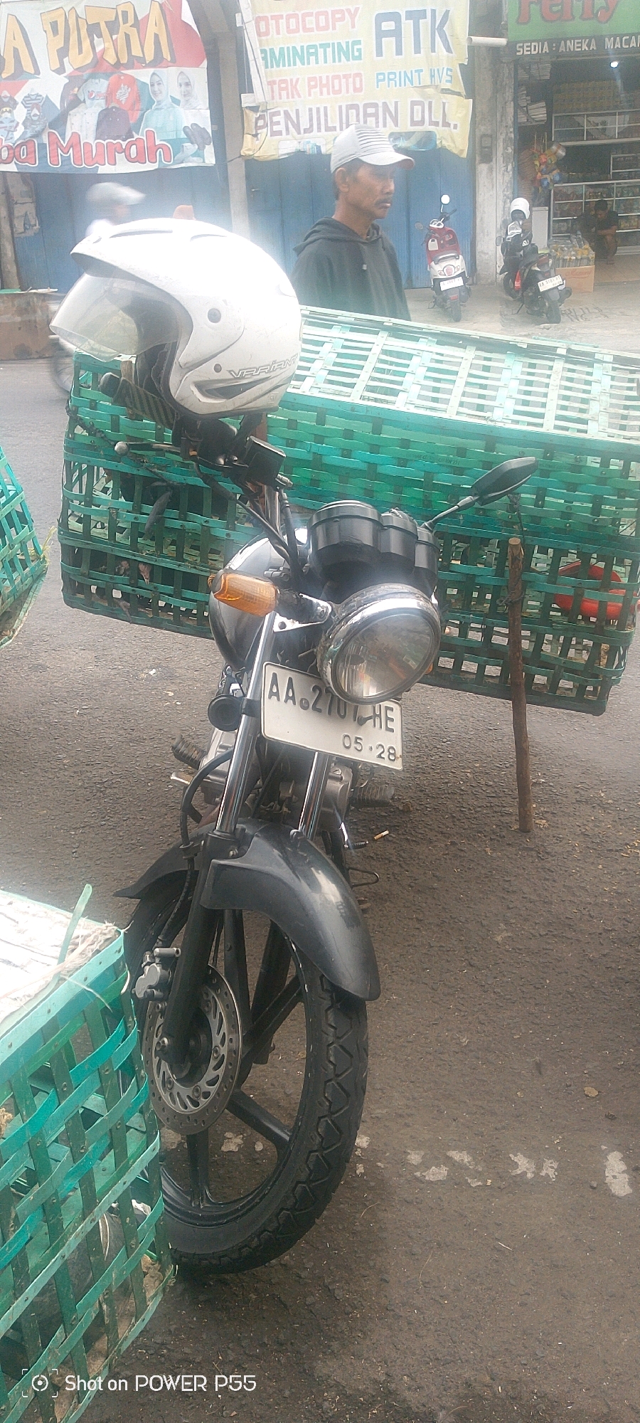 A motorcycle with a white helmet on its handlebars is parked on an asphalt road. The motorcycle has a license plate that reads "AA. 2701 HE" and "05-28". The motorcycle is loaded with two large, green, woven baskets, one on each side, extending from the front to the rear. The baskets appear to be carrying goods. In the background, a man wearing a gray baseball cap and a dark hooded sweatshirt is standing and looking to his right. Behind him, there are storefronts with various signs, one of which advertises "OTOTOCOPY ATK". Several other motorcycles are parked in the background near the storefronts. The scene suggests a street-side commercial area, possibly a marketplace.