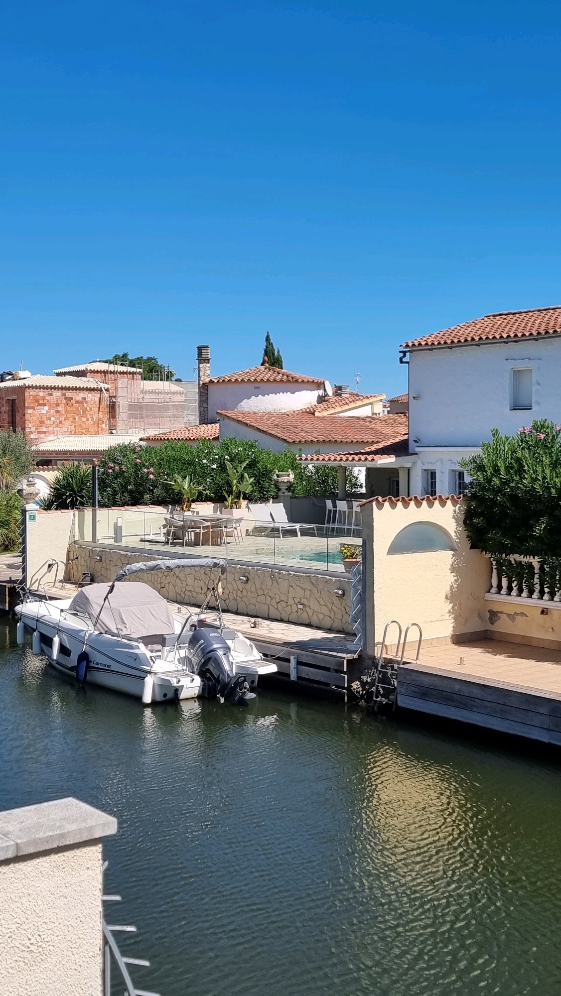 The image depicts a sunny day scene along a canal in Castelló d'Empúries, Spain, a town renowned for its picturesque canals and medieval architecture, often referred to as the "Venice of Empordà." In the foreground, two boats are moored at a wooden dock. The boat on the left is a white motorboat with a gray cover. The boat on the right is a smaller white motorboat with a prominent black Yamaha outboard motor. The canal water is dark and reflects the surrounding environment.
Behind the boats, a stone-walled terrace overlooks the canal, featuring a swimming pool with lounge chairs and a dining table with chairs. Greenery, including bushes and trees, is visible behind the terrace. Several residential buildings with terracotta tiled roofs are situated in the background. One building on the far left is under construction, with exposed brickwork and scaffolding. Another building to the right is a white two-story structure with a prominent window and a small balcony. The sky is clear and blue.
