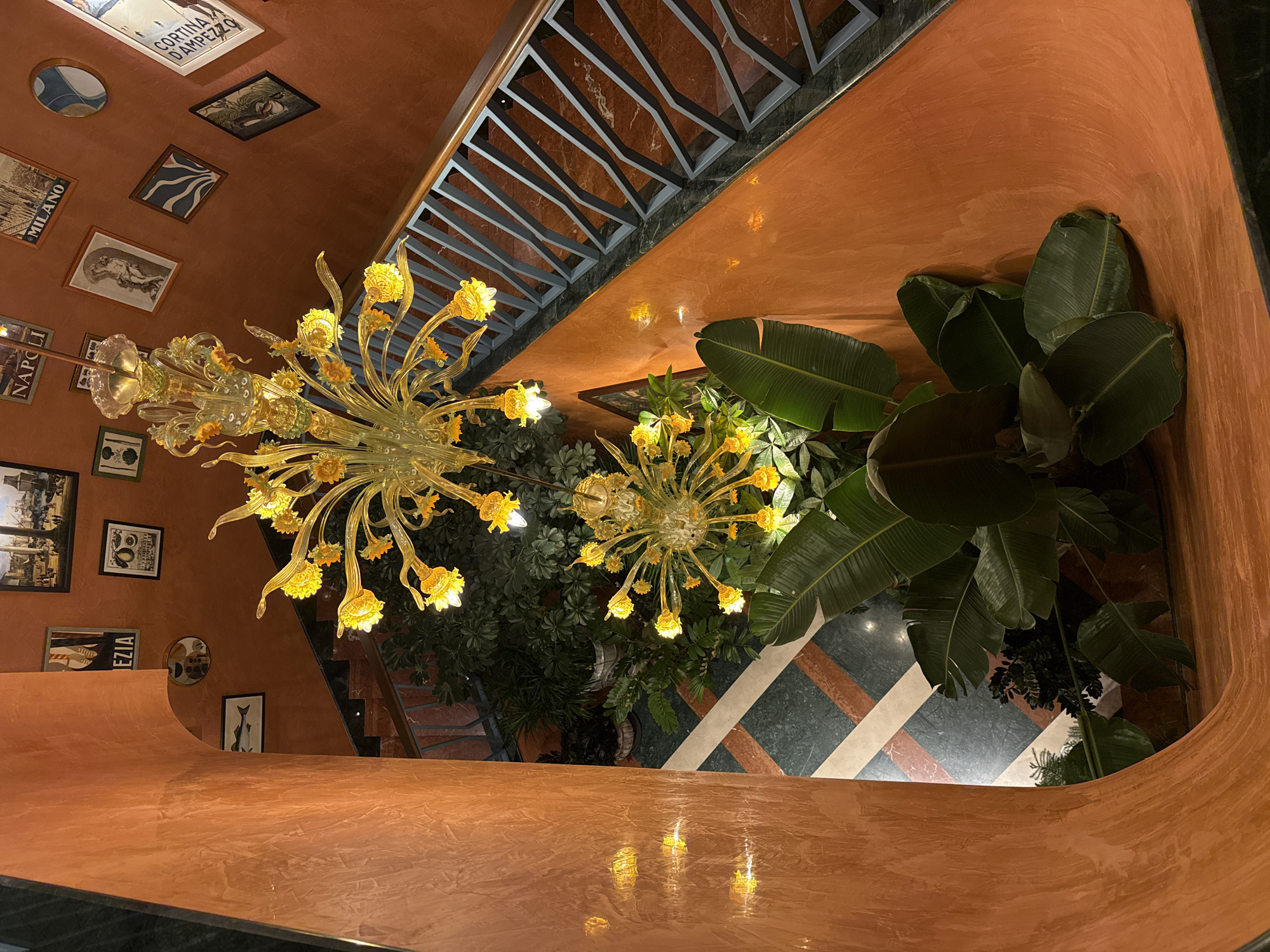 The image shows a dimly lit interior staircase with an orange textured wall. Two ornate yellow glass chandeliers hang from the ceiling, illuminating the space. The staircase has a dark railing with a geometric pattern and marble steps. Lush green plants are visible in the atrium below the stairs. The walls are adorned with framed pictures and posters, including one that reads "Venezia" and another that appears to be a travel poster. The flooring in the atrium features a geometric pattern of green, white, and reddish-brown tiles. The scene appears to be an indoor public space, possibly a hotel lobby or a restaurant. The location is identified as Baku, Azerbaijan.