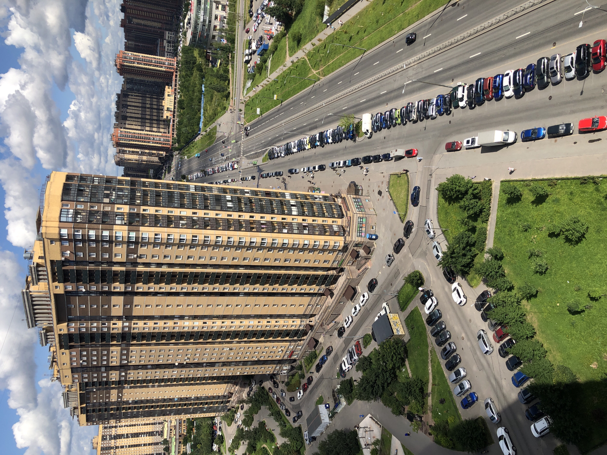 The image shows a long, multi-lane road running alongside a large, modern apartment building complex. The apartment buildings are tall, with many windows and balconies, and are predominantly yellow and brown in color. The road has a significant amount of parked cars lined up on both sides, as well as vehicles in motion. The scene appears to be in a developed urban or suburban area, with green spaces and other buildings visible in the background. The sky is partly cloudy.