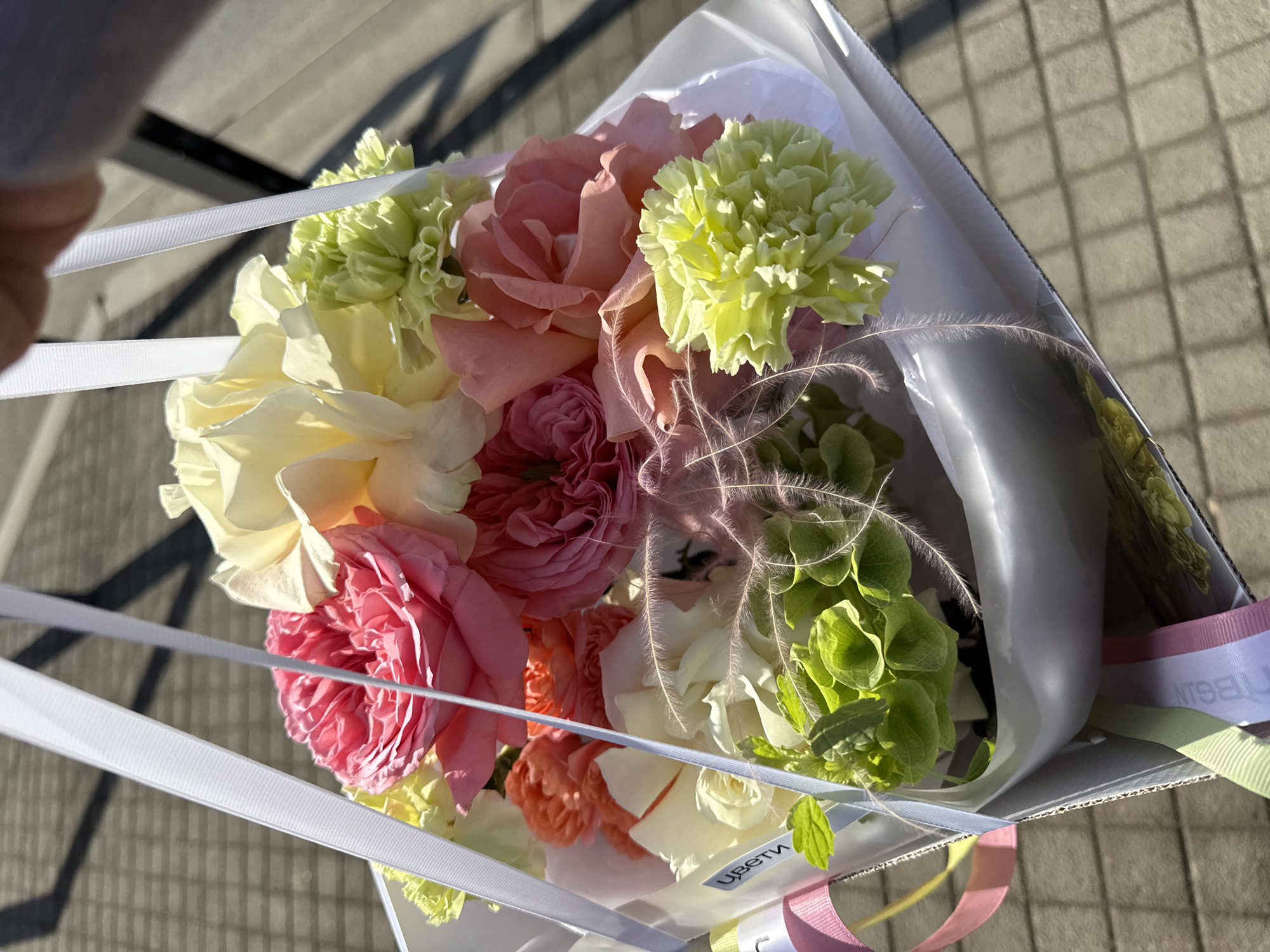 A bouquet of flowers is held up against a background of what appears to be a paved surface. The flowers include roses in shades of pink, peach, and white, as well as green carnations. The bouquet is wrapped in white paper and tied with white and multi-colored ribbons. The city of Perm, Russia is not discernible in the image.