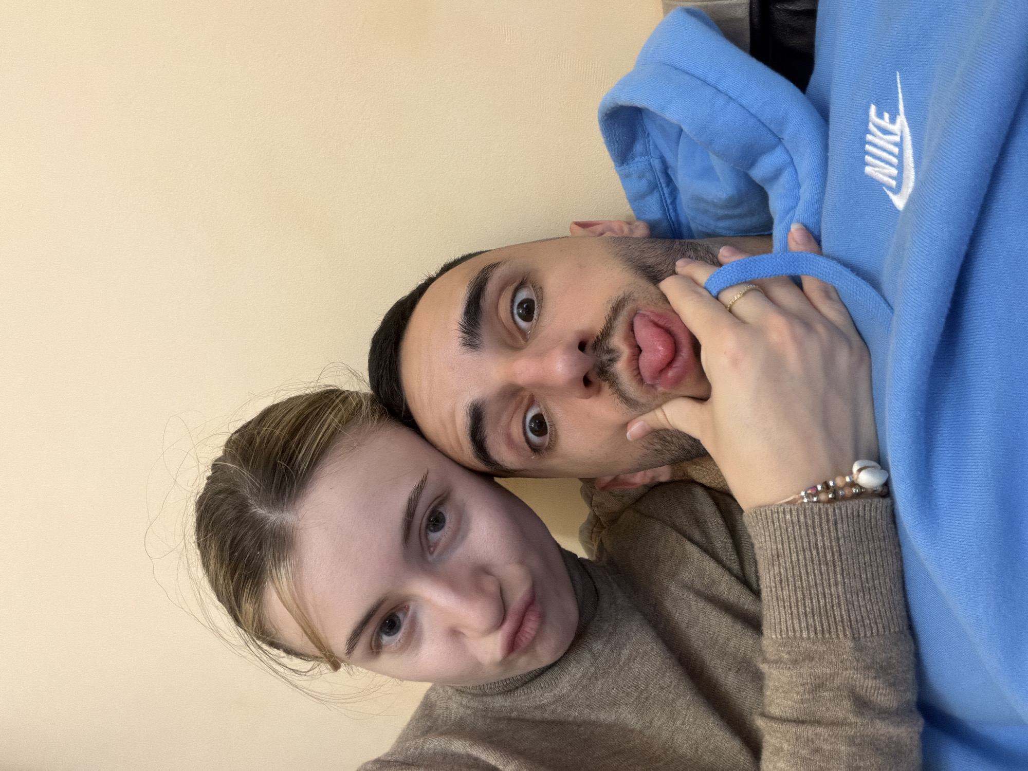 Two individuals are in close proximity to one another, likely a selfie taken indoors against a light yellow or beige wall. The man, positioned higher in the frame, is making a comical expression with his eyes wide open and his tongue sticking out. He has short dark hair and facial hair. A hand, likely belonging to the woman, is touching his face. The woman is positioned lower in the frame, with a pout on her lips. She has light-colored hair pulled back. She is wearing a brown turtleneck sweater, and there is a blue Nike hoodie visible in the background, partially covering the upper right of the frame. A bracelet is on the wrist of the hand touching the man's face. The location in Plovdiv, Bulgaria is not discernible from this photograph.