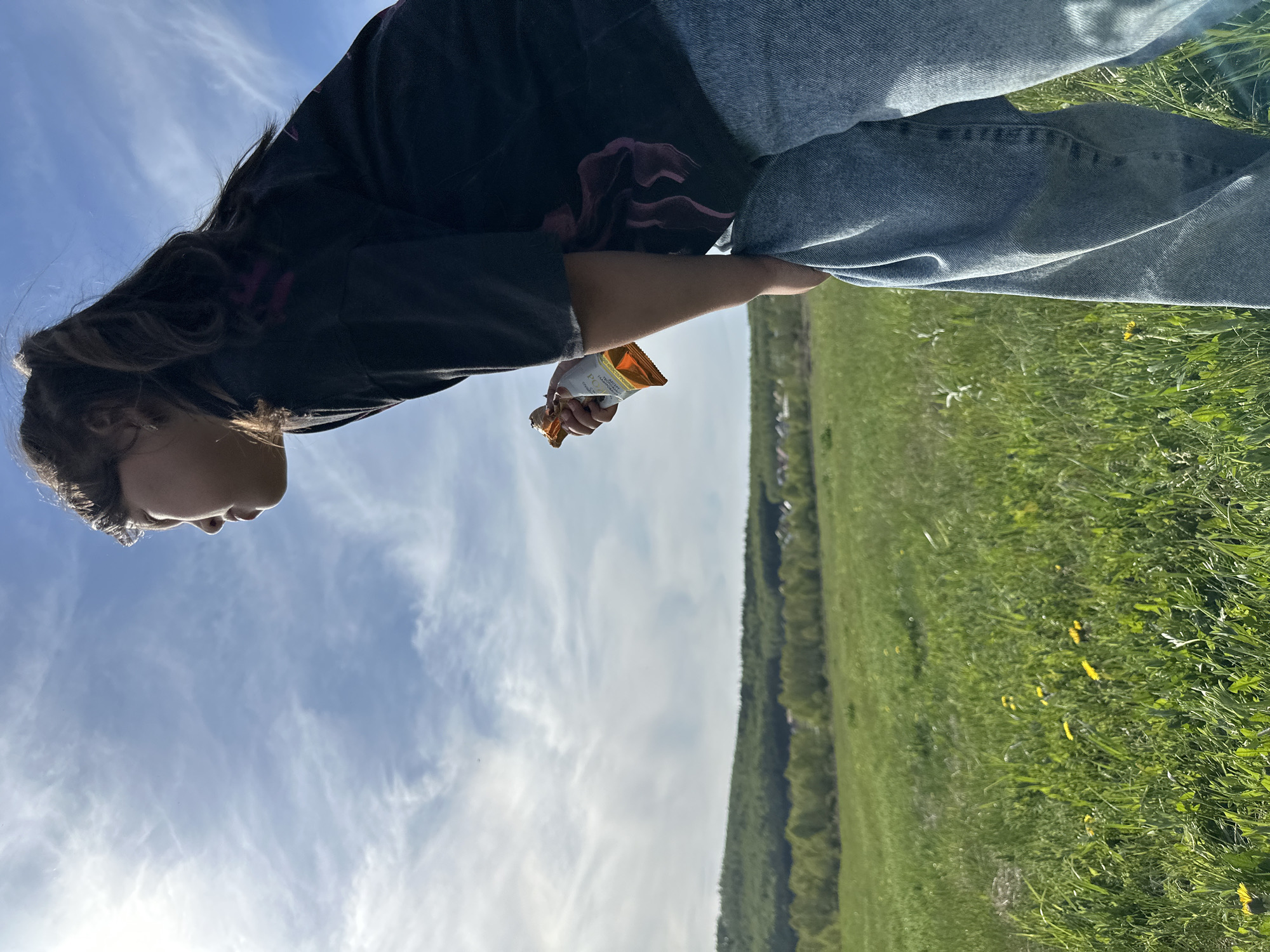 A person stands in a field, holding a partially eaten snack, against a backdrop of a blue sky with scattered clouds, a green field, and a treeline in the city of Unknown, Unknown. The person is wearing jeans and a dark-colored top. The snack appears to be in a gold wrapper. The field has patches of grass and some yellow flowers.