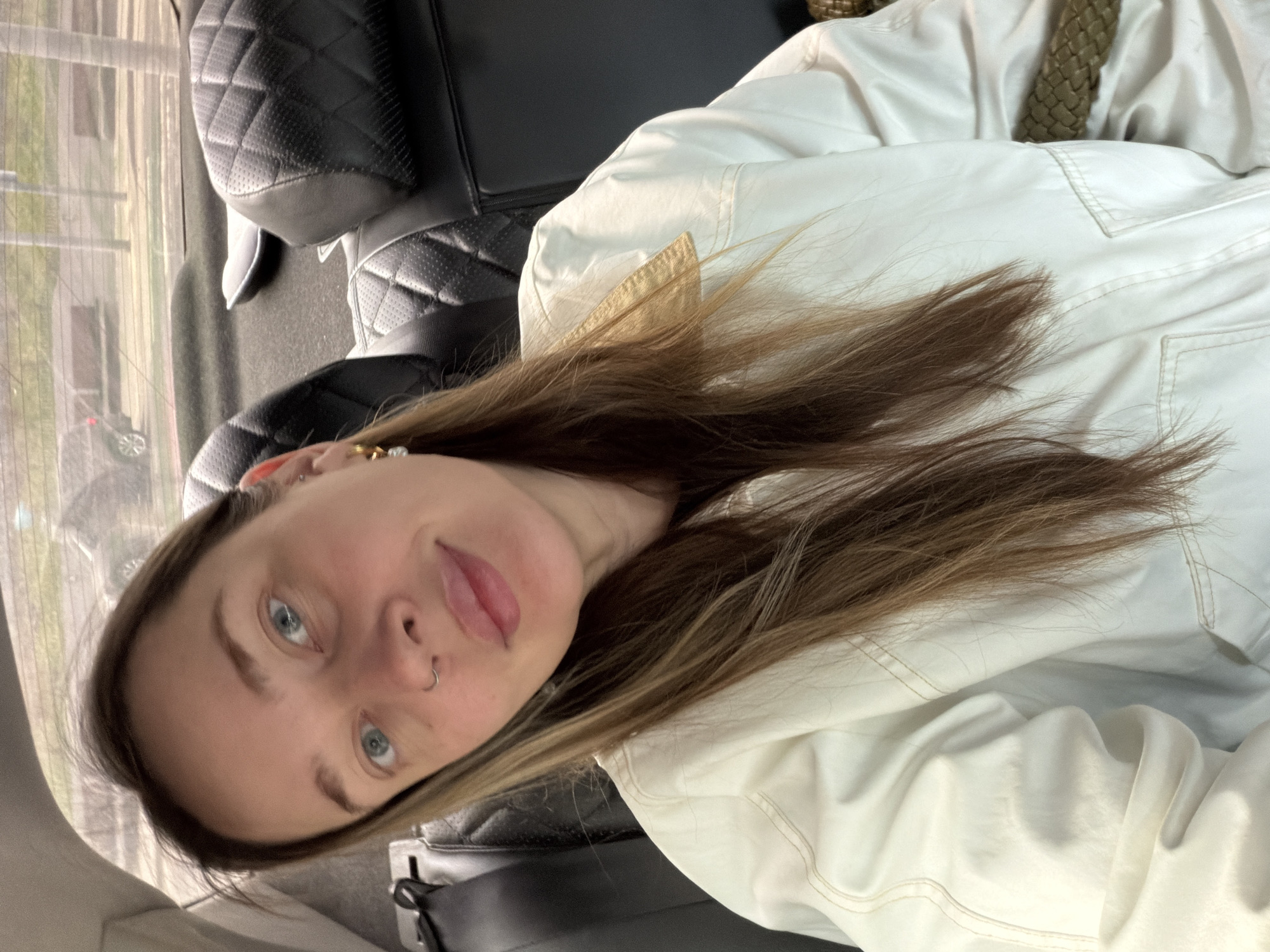 The image shows a person with fair skin, blue eyes, and light brown hair wearing a white jacket and a nose ring. The person is sitting in the back seat of a car with black leather seats. A view of the outside is visible through the car window, revealing an urban environment with buildings. The location is consistent with being in a vehicle within a city. The specific location of Перовское сельское поселение, Ukraine, cannot be confirmed solely from this image.