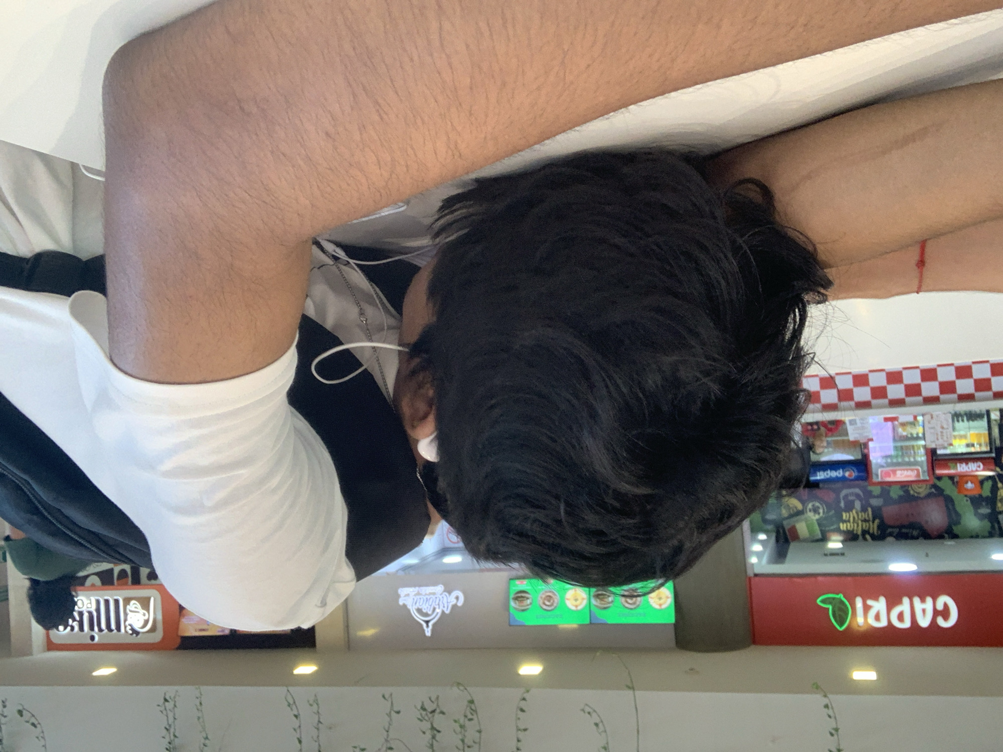 The image displays a person from behind, seated or leaning in an indoor commercial setting, likely a food court or mall in Pampatar, Venezuela. The individual has dark, wavy hair and is wearing a white t-shirt with a dark jacket or hoodie. White wired earphones are visible around their neck and in their ears, suggesting they are listening to something. Their arms, covered in fine hair, rest on a bright white surface, possibly a table or counter. A subtle red string bracelet adorns their right wrist.
The background features various illuminated signs and retail displays. On the left, parts of a "Minimarket" sign are visible, along with a sign for "Abdian Italian Pasta." Towards the center and right, a distinct red and white checkered pattern, reminiscent of a diner or pizza parlor, is present above display refrigerators. These refrigerators feature "Pepsi" and "Coca-Cola" branding. Below them, a prominent red sign with a green leaf logo clearly spells out "CAPRI" in white letters. Green digital menu screens showcasing food items can also be seen. The area below the signs displays decorative green, vine-like patterns, possibly on a wall or floor. The lighting suggests an active indoor commercial environment.