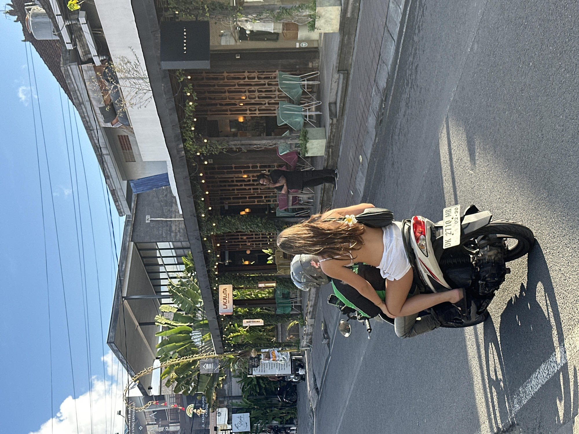 In North Kuta, Indonesia, two individuals are riding a scooter on a paved street. The person in the front is wearing a helmet, and the person in the back, identified as female, is wearing a white bikini top and white shorts, with a black purse slung over their shoulder. The scooter's license plate reads "DK 2710 MR". The background features a street lined with businesses, including "LACALITA" and "LOVEJOY", with outdoor seating areas and lush greenery. The sky is blue with some clouds, and sunlight casts shadows on the road.
