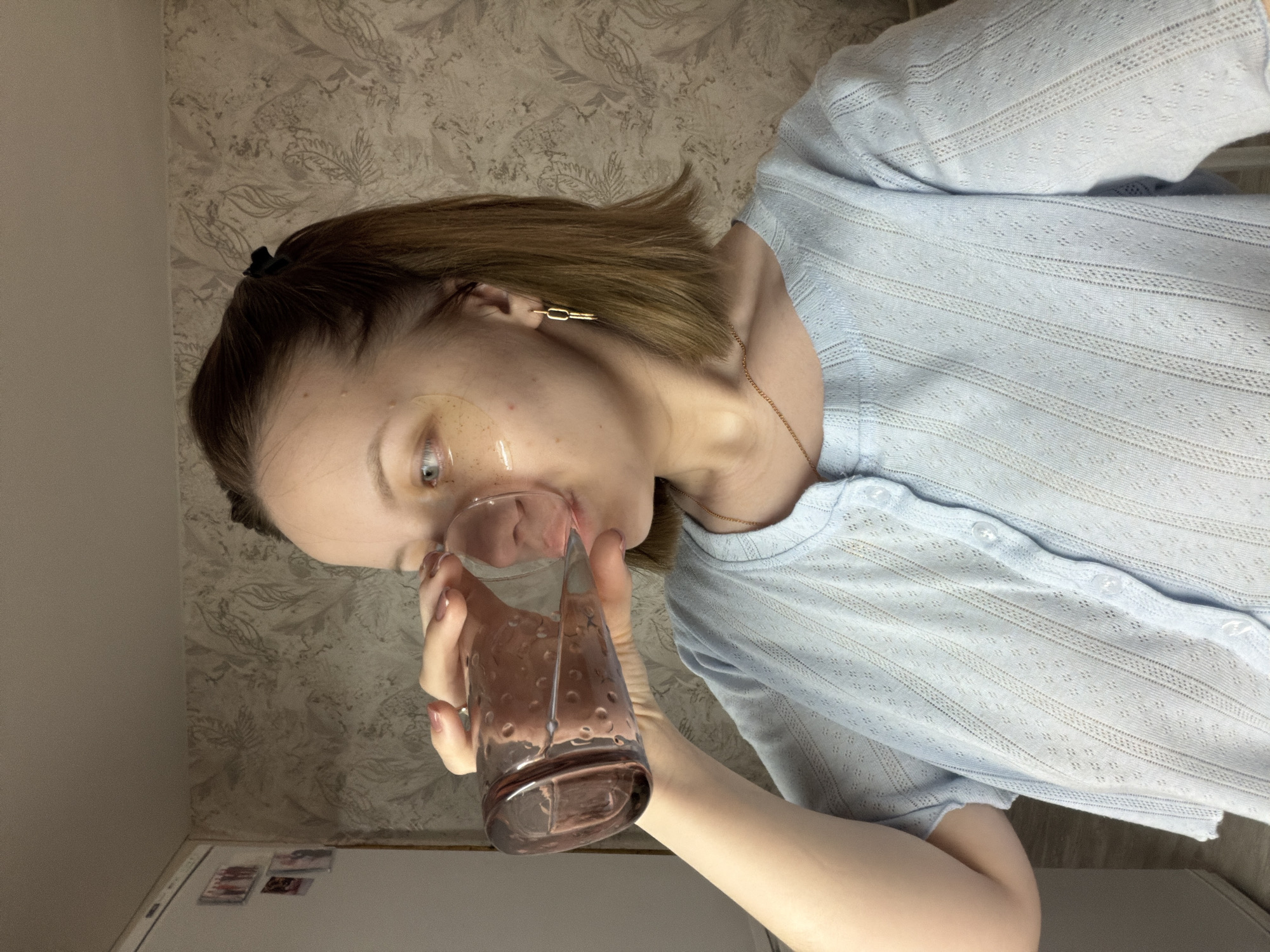A young person with light brown, shoulder-length hair is depicted indoors, holding a textured, translucent pinkish-purple glass to their mouth. They appear to be drinking or about to drink. Translucent, gel-like under-eye patches are visible beneath their eyes, suggesting a beauty or self-care routine. Their hair is partially pulled back with a dark scrunchie, and they wear a gold-colored chain earring on one ear and a thin gold necklace. They are dressed in a light blue, sheer, button-up shirt with an intricate patterned knit design. The background features patterned wallpaper with a leafy motif in muted grey and beige tones. Below the wallpaper, a white cabinet or appliance, possibly a refrigerator, is visible with several small magnets or photos attached. The lighting in the room is artificial. No discernable text is visible in the image. The scene is set in Reshetnikovo, Russia.