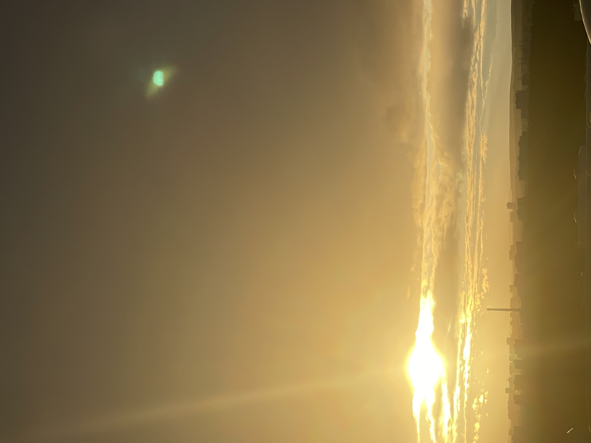 The image depicts a city skyline at sunset or sunrise, casting a warm, golden glow over the scene. The sun, a bright orb with visible rays, is partially obscured by clouds, creating a dramatic and atmospheric effect. A faint green lens flare is visible in the upper right quadrant of the sky. Below the sky, a silhouetted cityscape stretches across the horizon, with various buildings of different heights, including a prominent smokestack or tower. The foreground is also largely silhouetted, suggesting a higher vantage point from which the photograph was taken. The overall impression is one of a serene, urban landscape bathed in the soft light of dawn or dusk. The image is consistent with a view of the city of Perm, Russia, during these times of day.