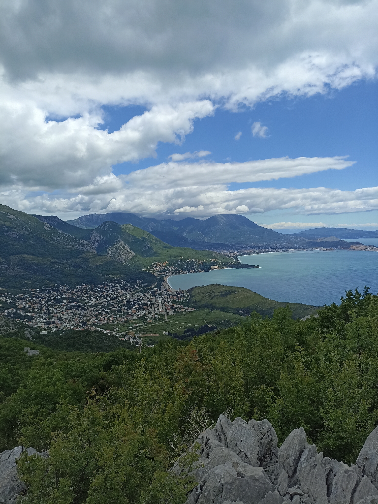 The image depicts a scenic view from a high vantage point near the city of Mišići, Montenegro. In the foreground, grey jagged rocks are visible, partially obscured by verdant green trees and bushes. Looking outwards, a coastal town sprawls along the shoreline, densely populated with buildings and structures. Behind the town rises a series of mountains, their slopes covered in varied shades of green vegetation. Further in the distance, a body of water, likely a bay or the sea, extends to the horizon. The sky above is filled with a mix of white cumulus clouds and patches of blue sky.