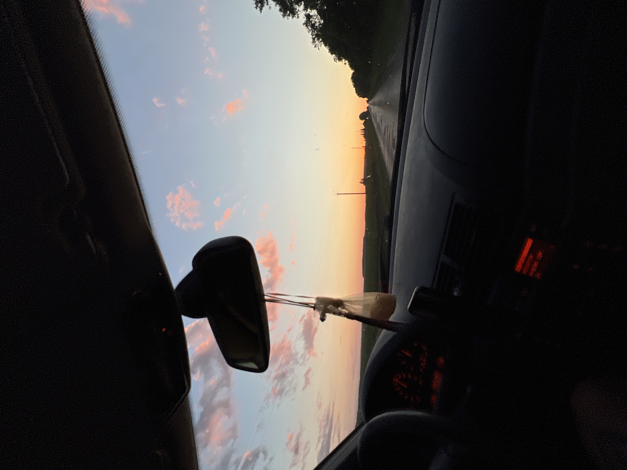 This media file depicts an interior view from a car as it travels along a road in Kypiachka, Ukraine. The scene is dominated by the sky, which displays a gradient of colors from pale blue at the top to a warm orange and pink near the horizon, indicating sunset or sunrise. Scattered pink clouds are visible against the blue sky.
The image is oriented sideways, with the dashboard and windshield on the left, and the right side of the car's interior on the right. A rearview mirror is prominently featured in the center-left of the frame, reflecting the sky. Hanging from the mirror are what appear to be air fresheners.
The road ahead is visible, receding into the distance and bordered by dark green trees on the left and a lighter green field on the right. A utility pole with wires can be seen along the roadside. The car's dashboard is partially visible on the right, with a red illuminated display that is difficult to read. Some of the dashboard instruments with red illumination are also visible.
The overall mood is peaceful and contemplative, capturing the beauty of a rural sunset or sunrise. There is no discernible activity involving people, but the presence of the car suggests travel. The lighting and colors suggest it is either early morning or late evening. No legible text is visible in the image.