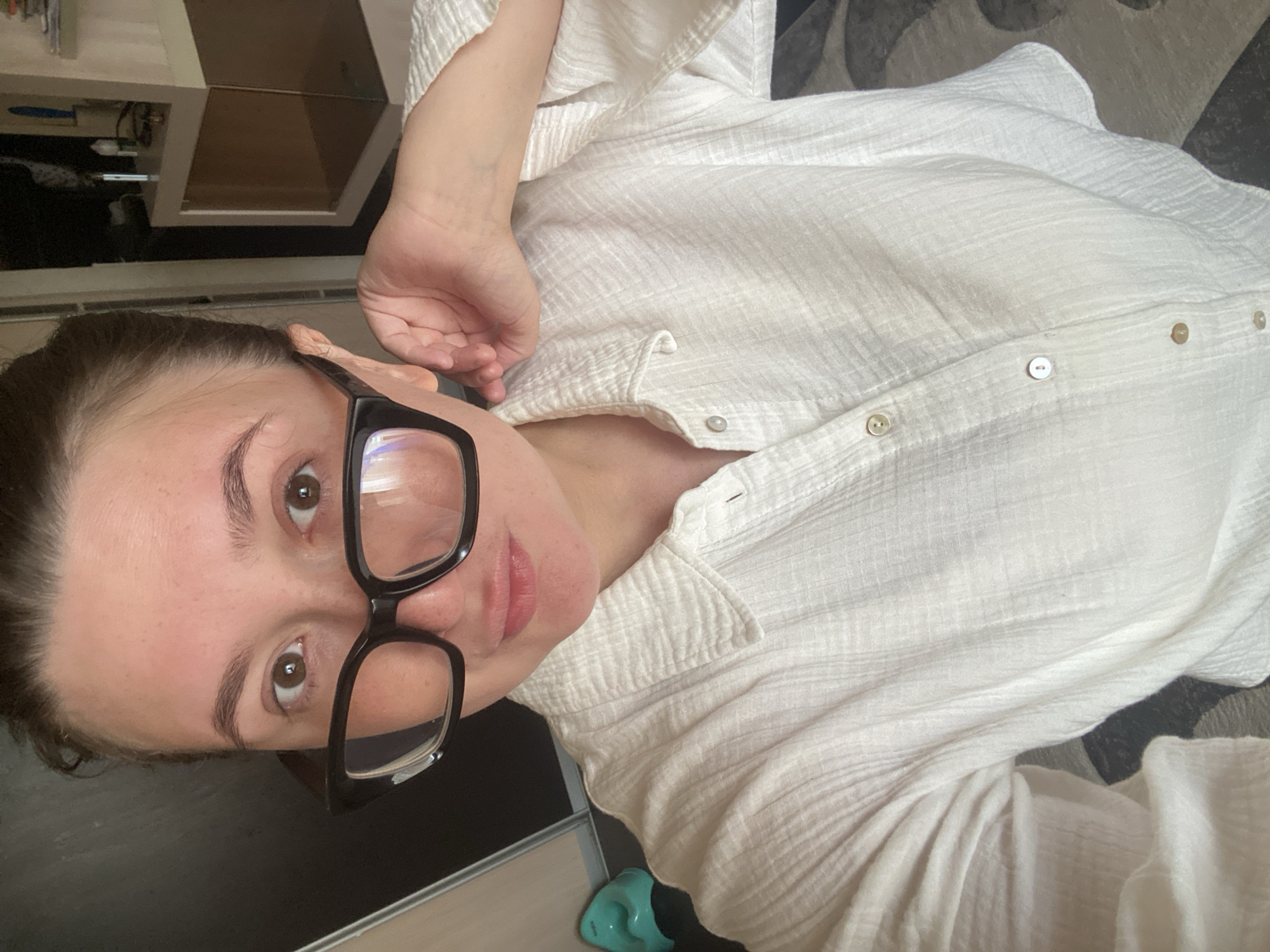 The image displays an upright, close-up indoor portrait of an adult individual, likely in Chaikovsky, Russia. The person, with fair skin, brown eyes, and dark eyebrows, looks directly at the camera with a calm, neutral expression and slightly parted lips. Their dark hair appears to be pulled back. They are wearing prominent black, thick-rimmed glasses.
The individual is dressed in a loose-fitting, white, textured button-down shirt, which appears to be made of a crinkled fabric like gauze. Several light-colored buttons are visible down the front of the shirt.
The background reveals a casual home environment. To the upper left, a light-toned piece of furniture, possibly a shelf or cabinet, is partially visible. A dark, flat surface, potentially a desk or table, is present in the lower left, where a small, light blue object can be seen. On the right side of the frame, a patterned rug or carpet in shades of grey and black is visible. The lighting is soft and ambient, typical of an indoor daytime setting. No text is visible in the image.
