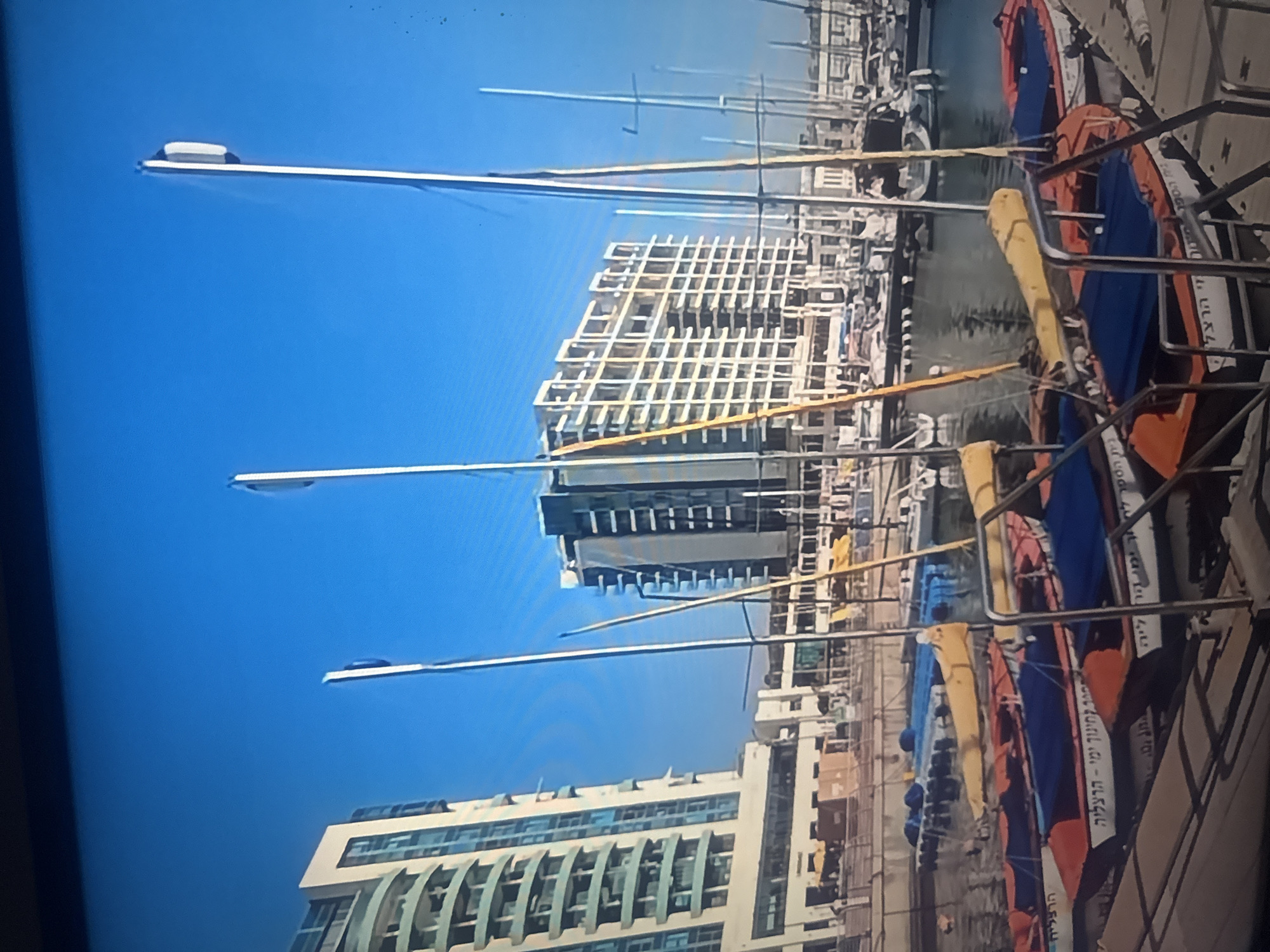 The image depicts a sunny day in the city of Herzliya, Israel, at the marina. In the foreground, several small, colorful boats are docked, some with blue coverings. Text in Hebrew is visible on the sides of the boats. Tall, modern buildings line the background, one on the left appearing to be residential with balconies, and another in the center with a grid-like facade. The sky is a clear, bright blue. Several tall mast poles, likely belonging to larger sailboats, rise above the smaller boats and buildings. The overall scene suggests a vibrant coastal city environment.