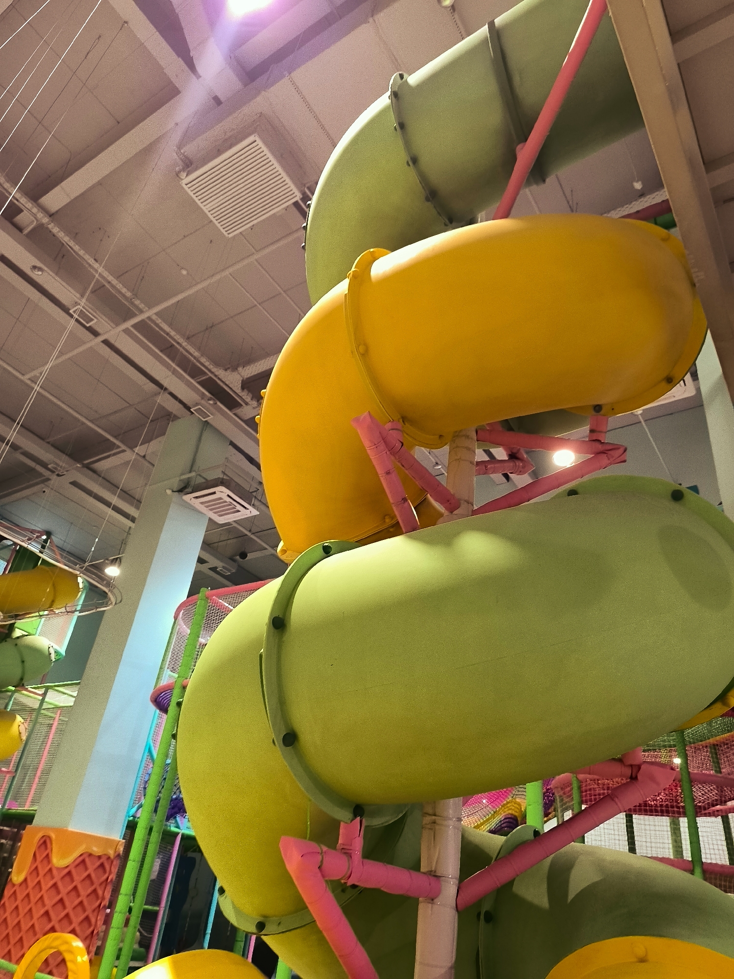 The image shows a brightly colored indoor playground with large, tubular slides in shades of green and yellow. These slides are supported by pink poles and metal structures, creating a multi-level play area. The ceiling is visible, featuring a ventilation grate and exposed light fixtures. The overall impression is one of a fun and energetic children's entertainment facility. There are no people visible in the image.
