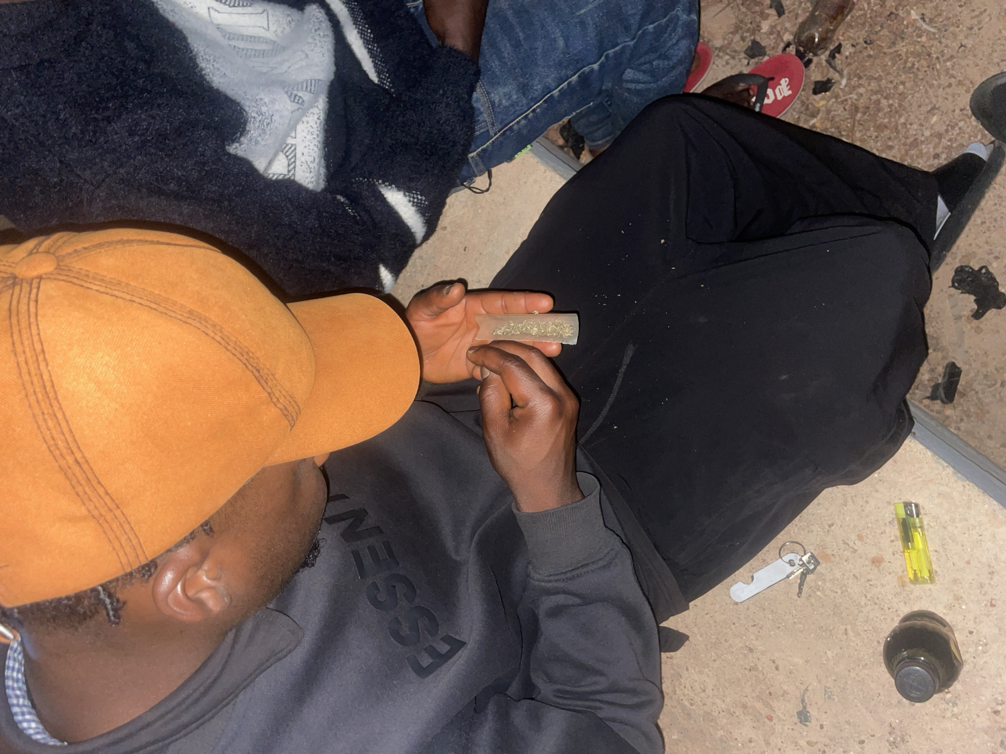 The image shows an overhead view of several individuals on a light-colored, possibly concrete, surface.
One individual, wearing an orange baseball cap and a dark sweatshirt with the word "ESSENTIALS" visible, is in the foreground. They are holding a rolling paper filled with a green leafy substance between their fingers.
To the side of this person are keys, a yellow lighter, and a dark bottle. Nearby, someone is wearing black sweatpants and black socks with white stripes. Another person is wearing blue jeans and red sandals.
No information is available regarding the location of Durimi, Nigeria or otherwise.