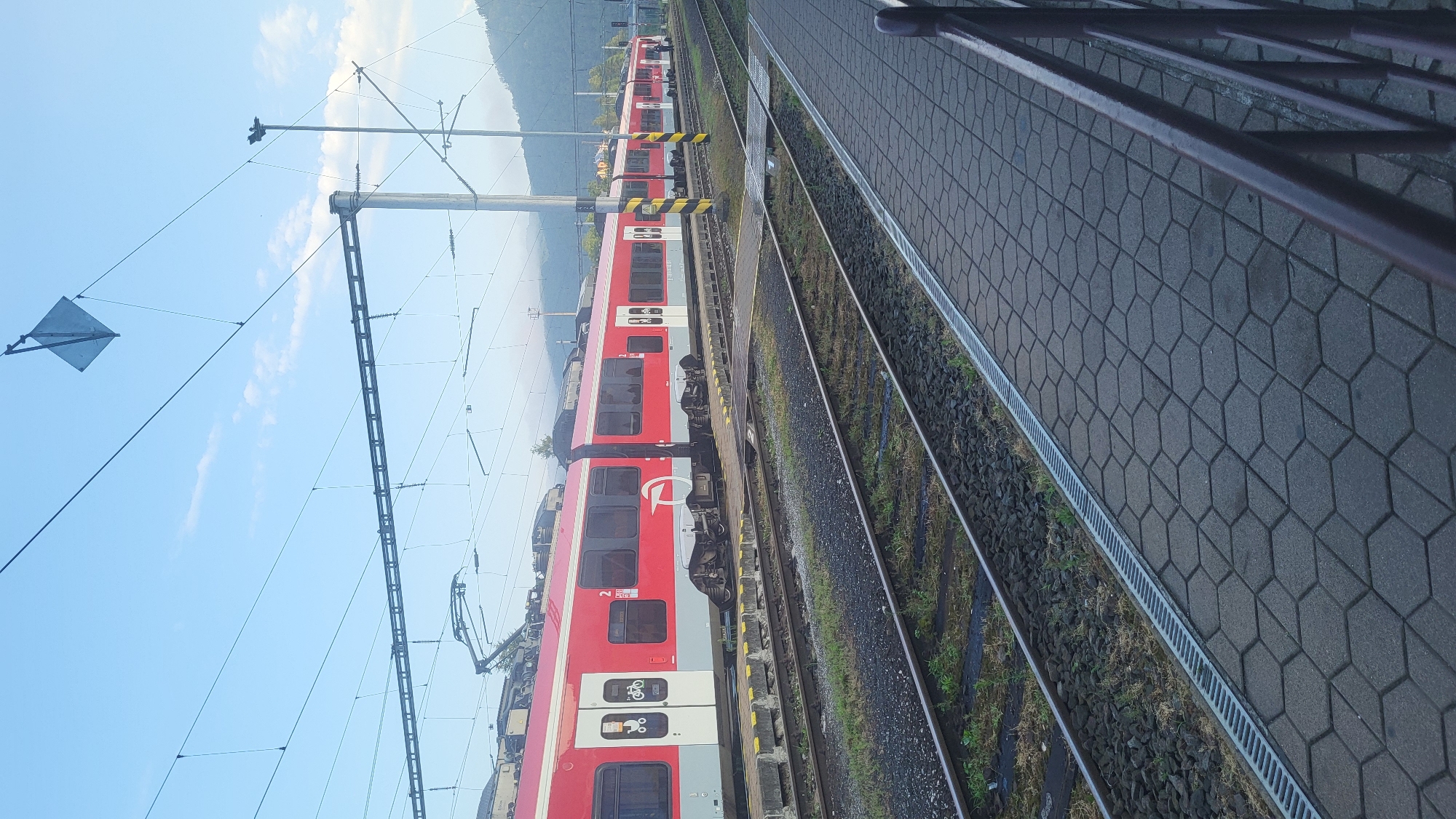 This is a photograph of a red and white train stopped at a train station in Kysak, Slovakia. The train is composed of several cars, and its livery features a white circular logo with a stylized "ÖBB" symbol. The train is on a track that runs parallel to a paved platform. The platform is made of hexagonal grey pavers and has a metal grate running along its edge. Adjacent to the platform are multiple sets of train tracks, with ballast and some vegetation visible between the rails. Overhead, a complex network of electrical wires for the train's power system is suspended from poles. In the background, hills covered in trees are visible under a blue sky with scattered clouds. The lighting suggests it is either early morning or late afternoon. There are no people clearly visible in the frame.