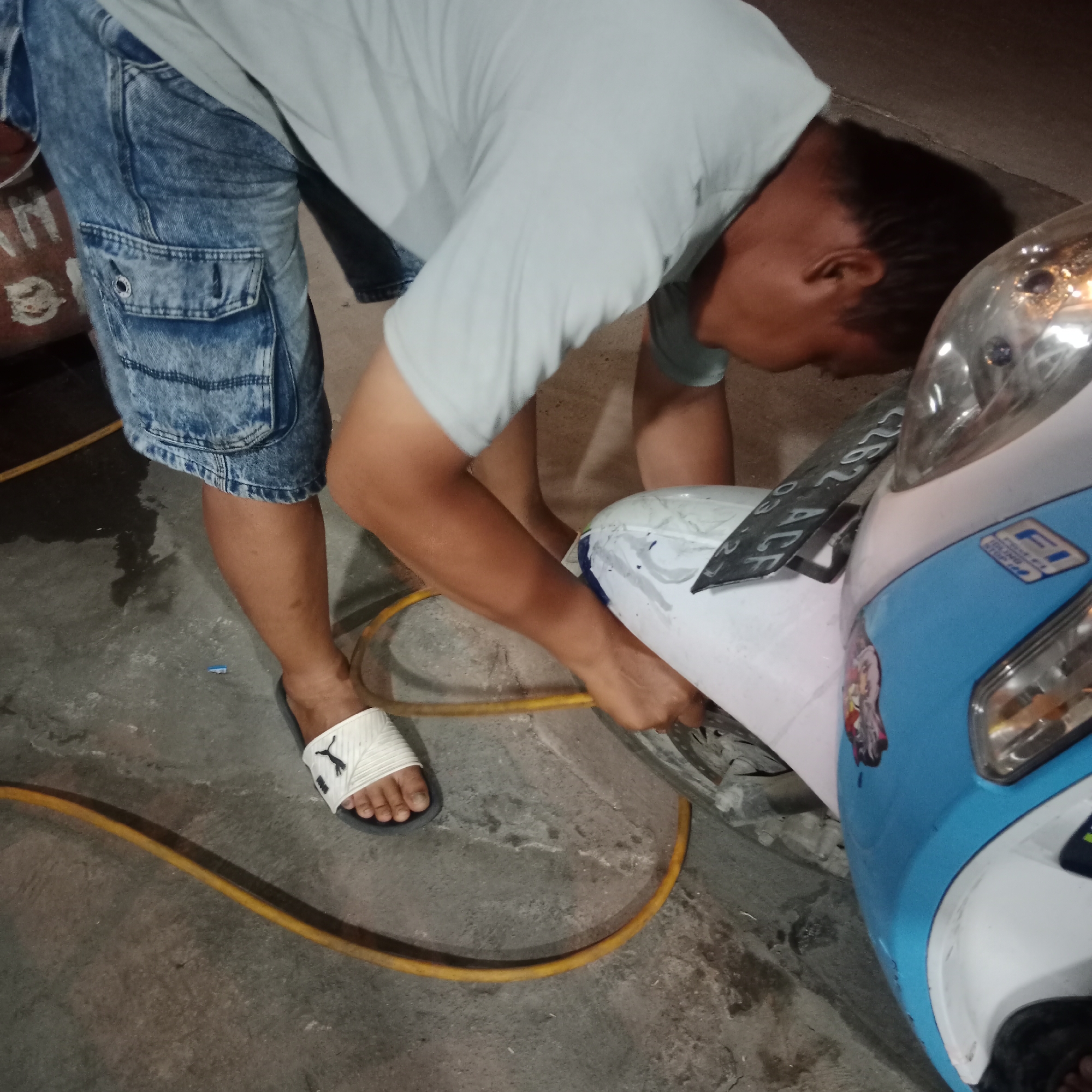 In Gubug, Indonesia, a person wearing denim cargo shorts and a light-colored t-shirt is shown leaning over a blue and white motorcycle. The person is holding a yellow hose, which appears to be connected to the motorcycle. The ground is a concrete surface with some wet patches. A license plate with the characters "3262 ACF" is visible on the motorcycle. The lighting suggests it is nighttime or a dimly lit area.