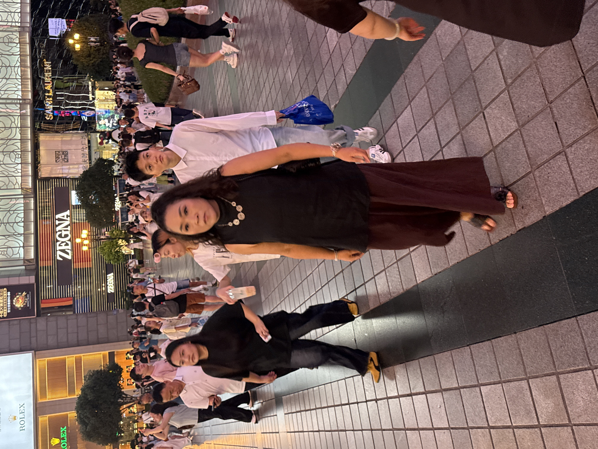 The image depicts a street scene in Yuzhong District, China, likely in the evening. Several people are walking on a tiled pedestrian walkway in front of a large building with illuminated signage for brands like ROLEX and ZEGNA, as well as "SAINT LAURENT". In the foreground, a woman in a black sleeveless top and a dark red skirt is walking towards the viewer, wearing a prominent necklace and sandals. To her left, another woman in a black top and dark jeans is walking away, also wearing sandals. A man in a white shirt and jeans is walking behind the woman in the skirt. Other individuals are visible in the background, some carrying shopping bags. The overall atmosphere suggests a bustling commercial area.