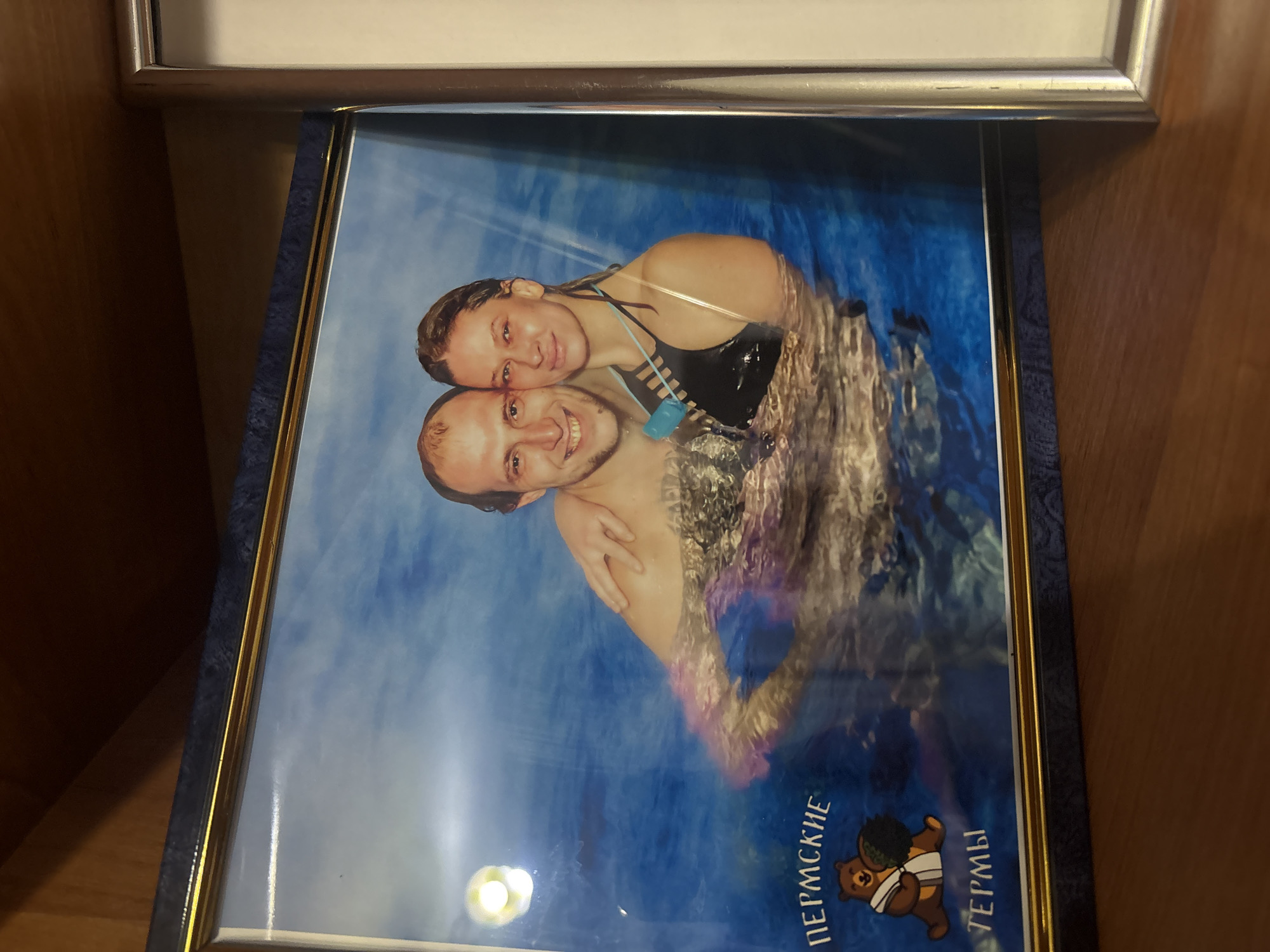The image is a framed photograph of a couple in a swimming pool. The photograph is tilted and shows the couple with their heads close together. The woman is wearing a black swimsuit, and the man has his arms around her. The photo is framed in a blue and gold frame. At the bottom of the image are the words "ПЕРМСКИЕ ТЕРМЫ" and a logo of a bear, indicating the photo was likely taken at the Permskie Termy water park in Perm, Russia.