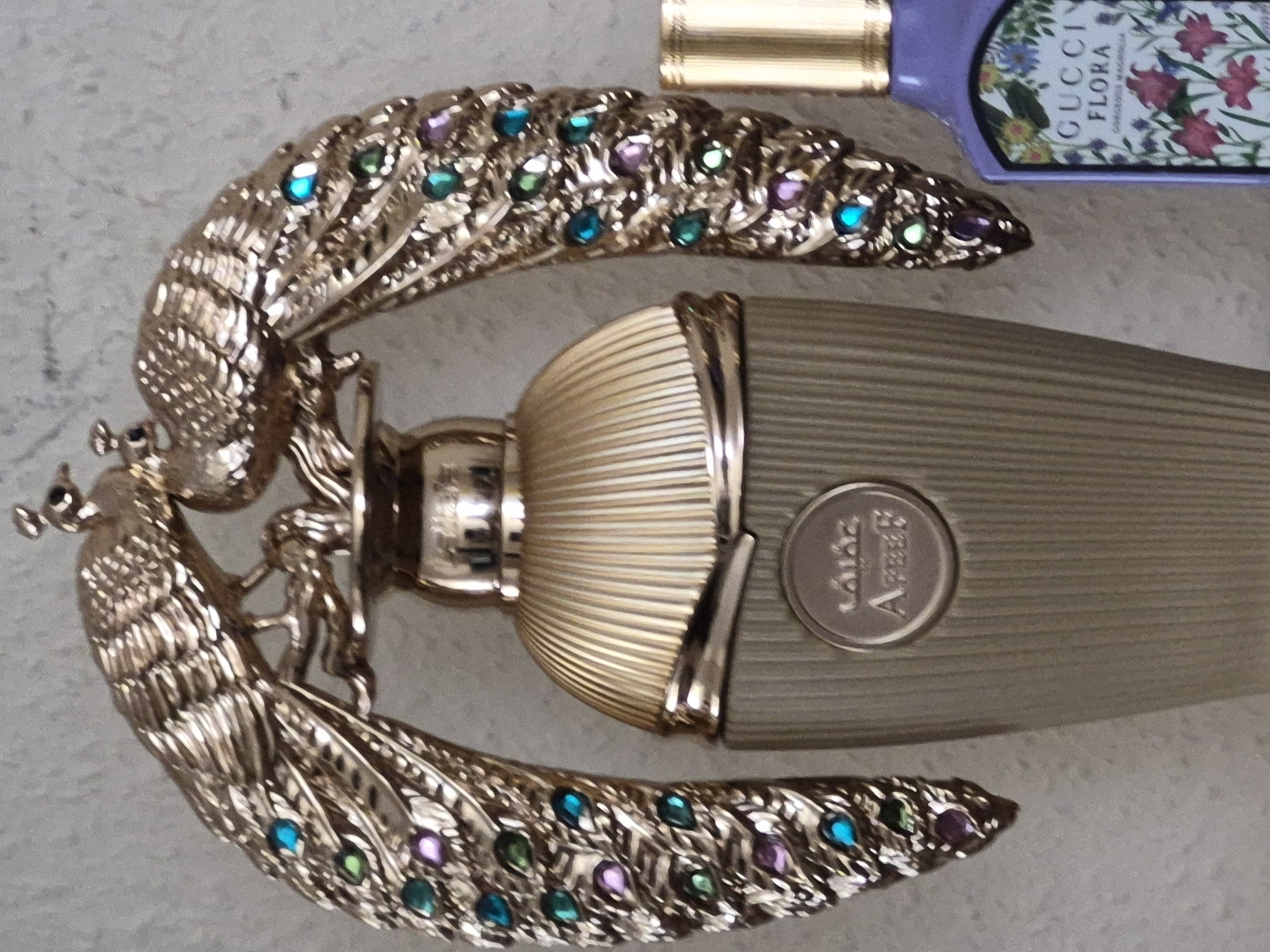 This media file is a close-up, overhead shot of a decorative perfume bottle with an ornate, gold-colored peacock-shaped cap. The cap is highly detailed, featuring the body and fanned tail of a peacock, adorned with small, colorful gemstones in shades of blue, green, and purple. There are also small figures of what appear to be humans or cherubs integrated into the peacock design. The body of the perfume bottle itself is a textured, metallic gold color with vertical ribbed detailing. To the right of the peacock-shaped cap, the body of a second perfume bottle is visible. This second bottle is clear and contains a pale purple liquid, with a label featuring floral imagery and text.
Visible text on the label of the second perfume bottle reads: "GUCCI FLORA." There is also a circular emblem on the body of the first perfume bottle with some text, but it is too blurry to be clearly deciphered. The overall scene appears to be a still life arrangement of perfume bottles, possibly set on a textured surface like a table or countertop. The lighting is somewhat diffused, suggesting an indoor setting. No specific time of day, weather conditions, or emotional cues are discernible from the image.