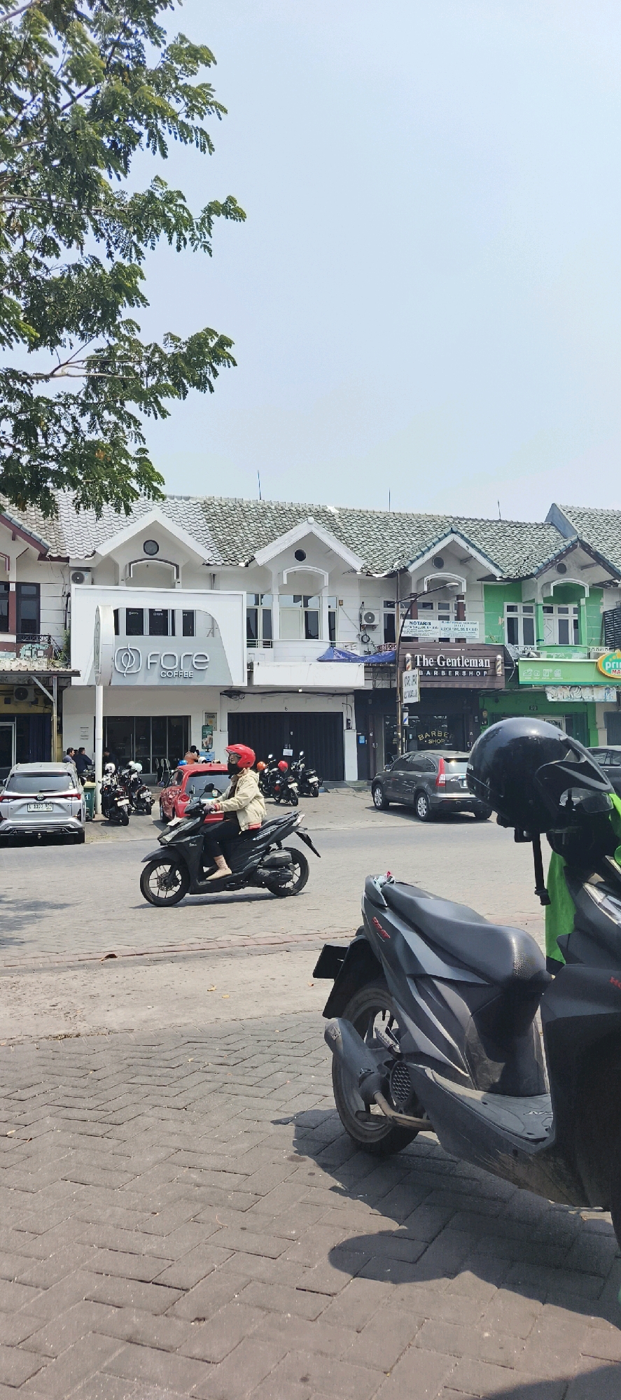 In Kureksari, Indonesia, a street scene unfolds under a clear sky. In the foreground, a black motorcycle with a visible exhaust pipe is parked, casting a shadow on the herringbone-patterned brick pavement. Behind it, another motorcycle, also black, is partially visible, with a green bag draped over its handlebars.
The middle ground features a paved road with a person riding a black scooter, wearing a red helmet and a light-colored jacket. Several other parked motorcycles are visible to the left of the scooter rider, as well as a silver car. Red and white buildings with green tiled roofs line the street. One building prominently displays a sign for "Fore Coffee". Another building to its right has a sign for "The Gentleman Barbershop". A third building further to the right has green accents and signage that is partially obscured. Trees with green foliage extend from the top left of the frame, partially obscuring the sky.