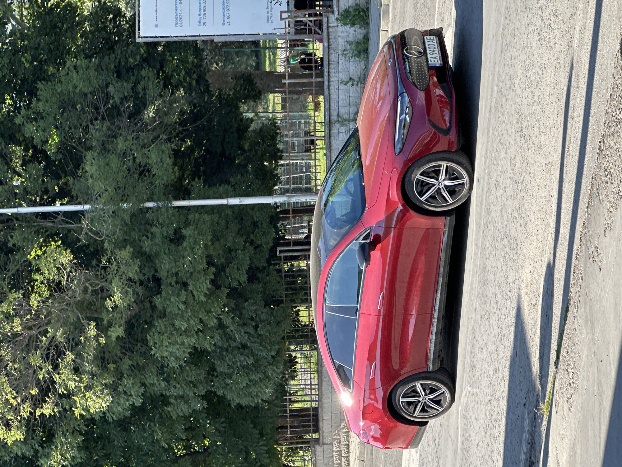 A red Mercedes-Benz car is parked on a concrete surface in what appears to be an urban or suburban setting. The car is viewed from the side, angled towards the viewer. Behind the car, lush green trees form a dense backdrop, suggesting a location with ample greenery. A metal fence is visible in the mid-ground, separating the car and the viewer from the trees and what might be a park or public space. The scene is bathed in sunlight, casting long shadows from the car and other unseen objects onto the ground, indicating it is likely late afternoon or early morning.
Visible text on a signpost near the car provides partial information. It includes what seems to be a website "www.veliko-tarnovo.bg", and financial or contractual details such as "Продължител 04/2024 г. - 04", "Общ бюджет 25 726 909.32", and "Финансиран 21 867 872.92". There is also text identifying an office location: "Офис: гр. Велик" and "ул. Поп Харито". The license plate of the car reads "EA 9400 AE". The overall impression is a bright, sunny day in a location that combines urban elements with natural scenery.