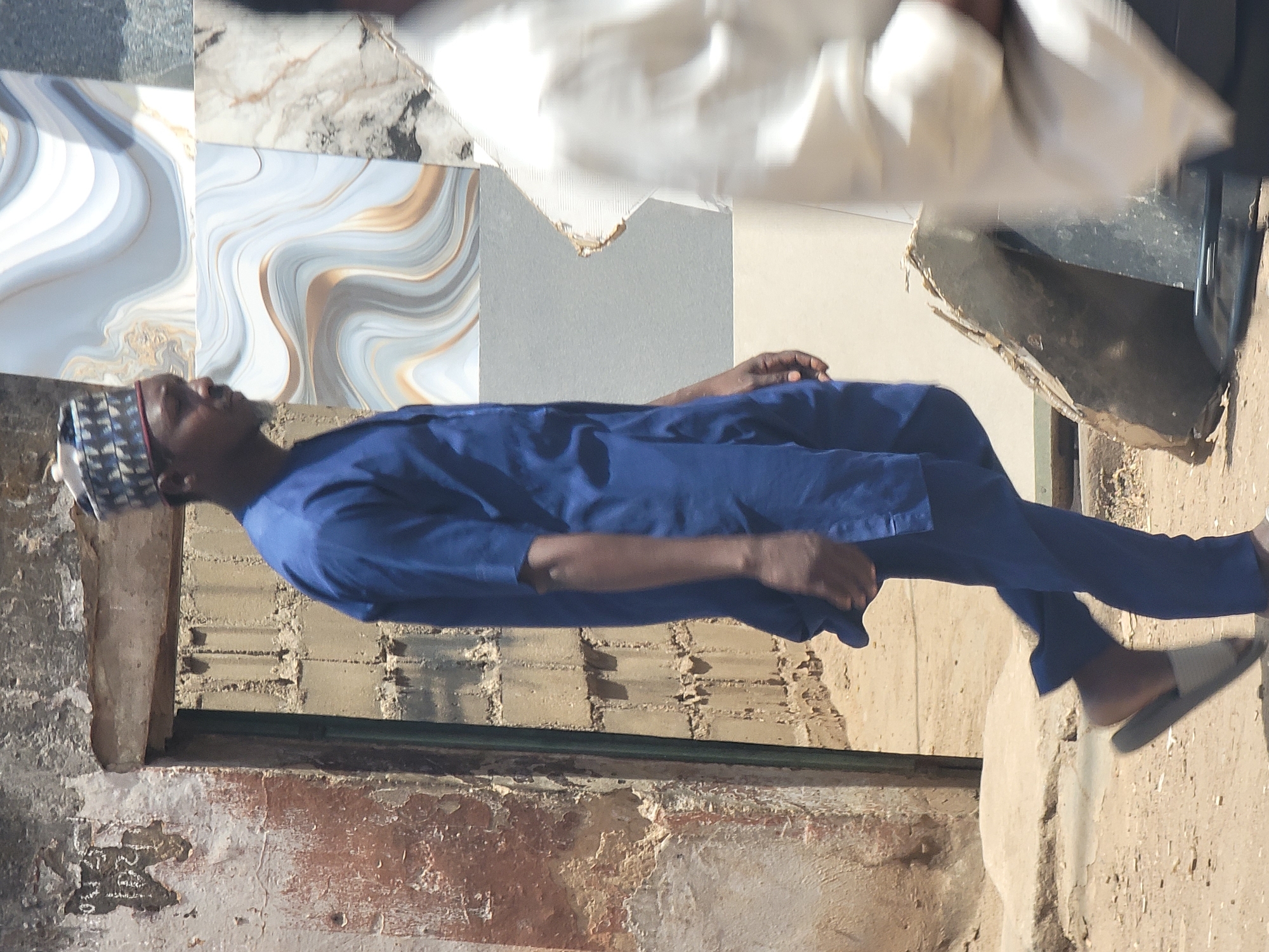 The image, oriented correctly, features a man standing in an outdoor or semi-outdoor setting in Maiburiji, Nigeria, under bright daylight. The man, with dark skin, is positioned slightly to the left of the frame, looking upwards and slightly to his left with a neutral expression.
He is dressed in a royal blue long-sleeved tunic over matching blue trousers. On his head, he wears a patterned cap with a geometric design in white and dark blue/black. He has grey slide sandals on his feet, and his hands rest casually by his sides.
The background behind him is textured and varied. To his upper left and directly behind his head, two large square tiles with a swirled pattern in shades of blue, white, and golden-brown are visible, resembling polished stone. Below these tiles, there is a plain gray wall section. Further down and spanning the lower left of the frame is a rough, worn wall or foundation made of exposed bricks and peeling plaster, displaying earthy tones of brown, white, and reddish-orange. To the man's right, a dark, vertical structure, possibly a doorframe or pillar, is partially visible. The ground beneath him is uneven, sandy-colored earth, casting distinct shadows that suggest strong sunlight. In the far upper right background, a blurred, light-colored object or fabric is faintly discernible. No discernible text is visible anywhere in the image.